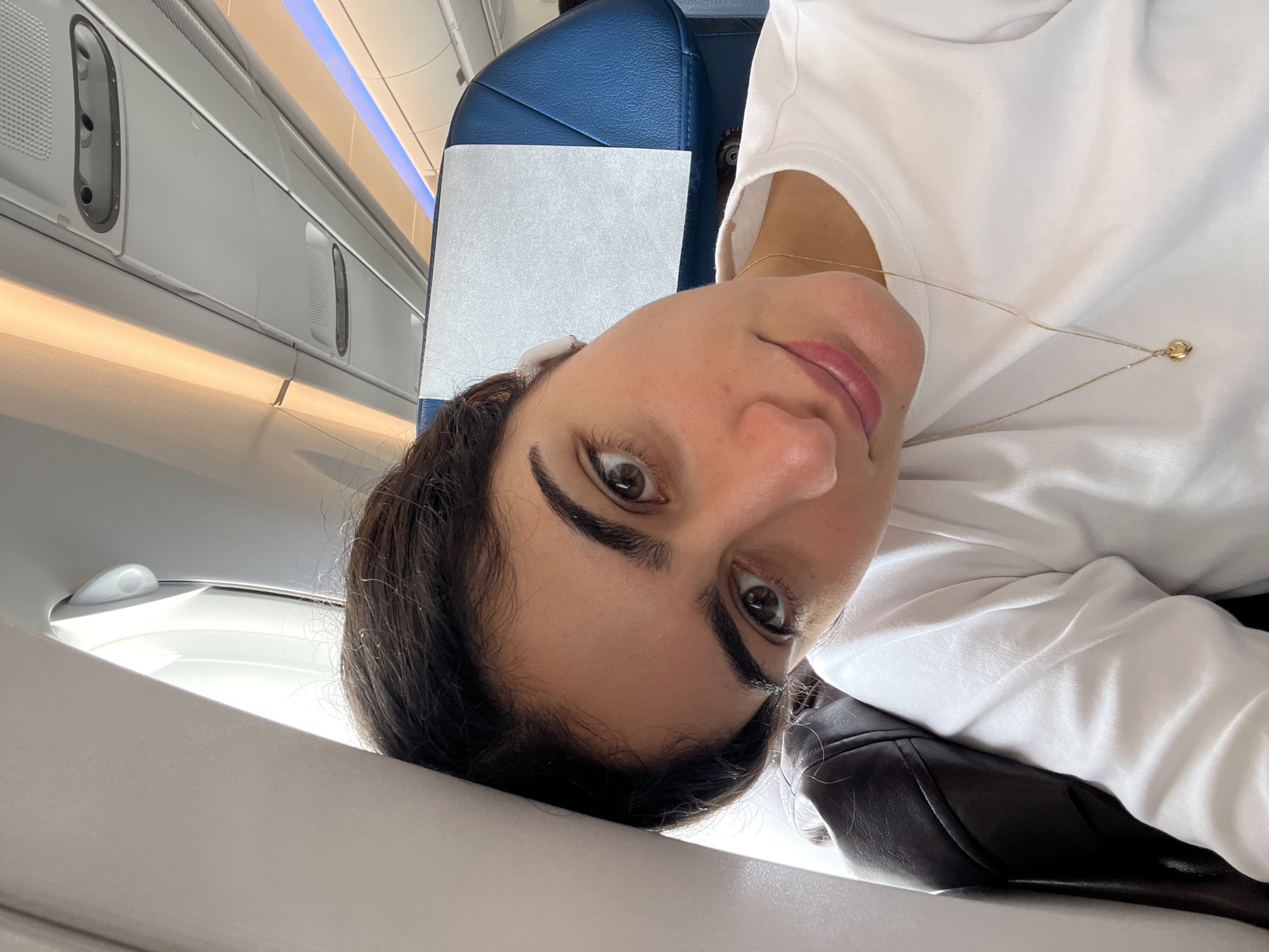 A woman is in the cabin of an airplane. The plane appears to be over Orly, France based on the location in the metadata. The woman is wearing a white t-shirt and a gold necklace. She has dark hair and brown eyes. She is looking at the camera. The airplane cabin has overhead lights and a blue seat with a white headrest.