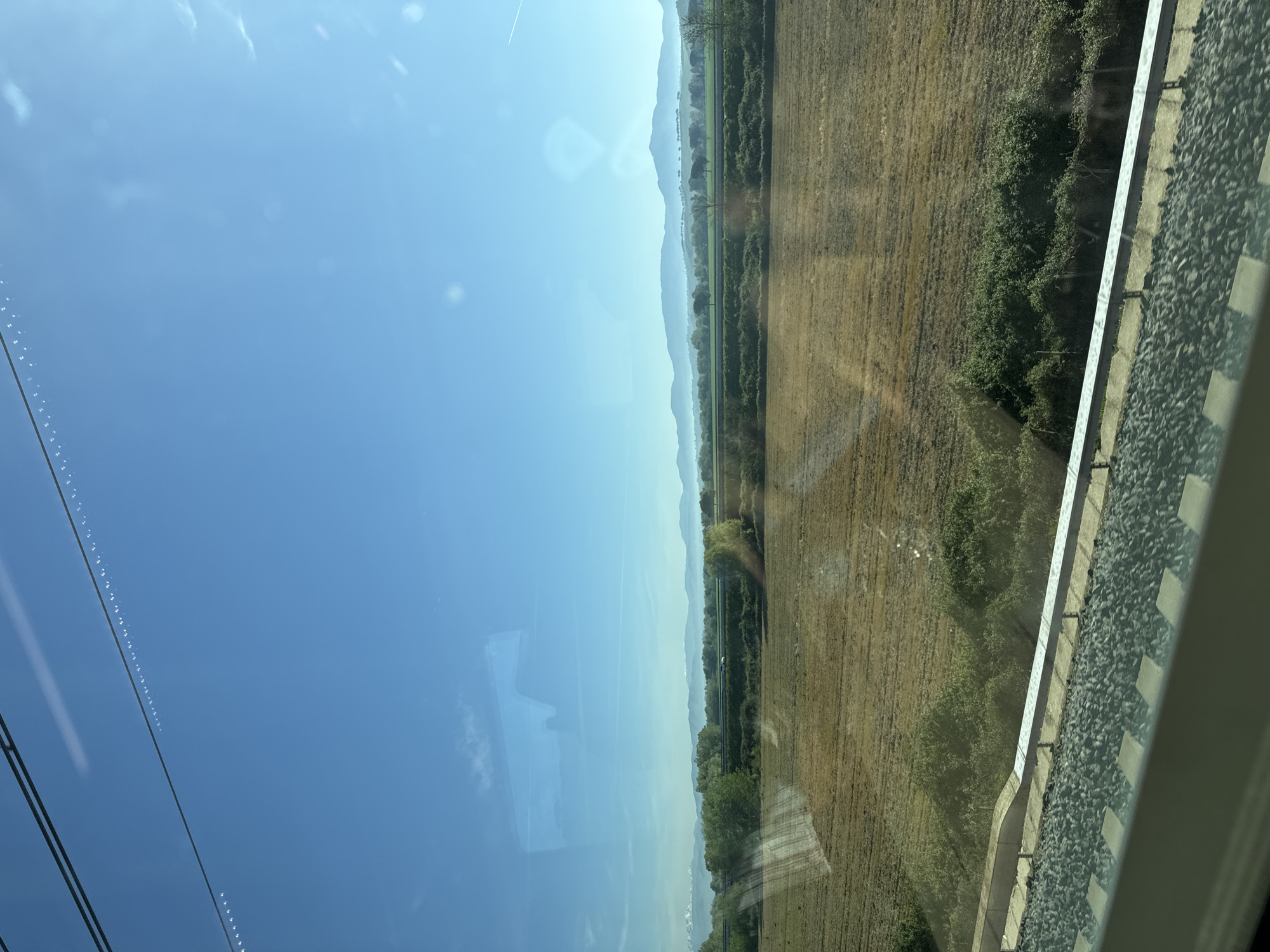 A horizontal outdoor shot taken through a train window on a bright day. The sky is a gradient of light blue, with some faint clouds visible. In the distance are hazy, layered mountains. A field of dried grass stretches out next to a line of lush green trees and bushes. To the right of the field, a railway track with grey gravel and white block markers is visible. The image appears to be taken from a moving train.
The location cannot be definitively determined as Rome, Italy, based on the visual content of the image alone.