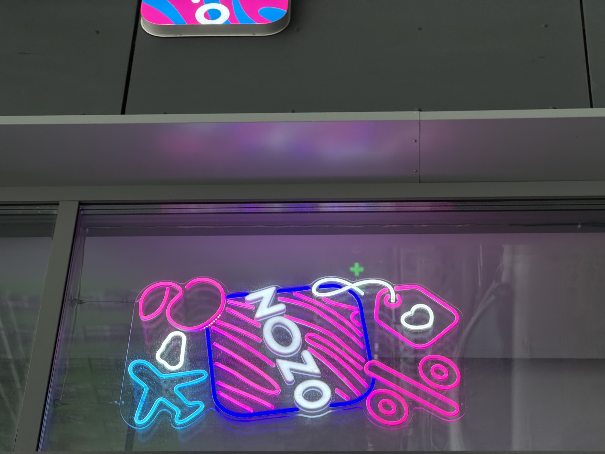 A close-up, vertical shot features a neon sign in the window of a building, reflecting ambient light and the urban environment outside. The sign is primarily pink and blue, with white accents. It depicts various symbols: a light blue airplane, a white cloud, a rectangle with pink diagonal stripes containing the white text "OZON," a pink percentage sign, and a pink tag with a white heart outline. The sign is illuminated and positioned behind a window. To the right of the window, a portion of a grey building facade is visible, along with another neon sign that is partially obscured and predominantly pink and blue, with white lettering that appears to start with "O." The setting is ambiguous, but the presence of a store window suggests a commercial area.