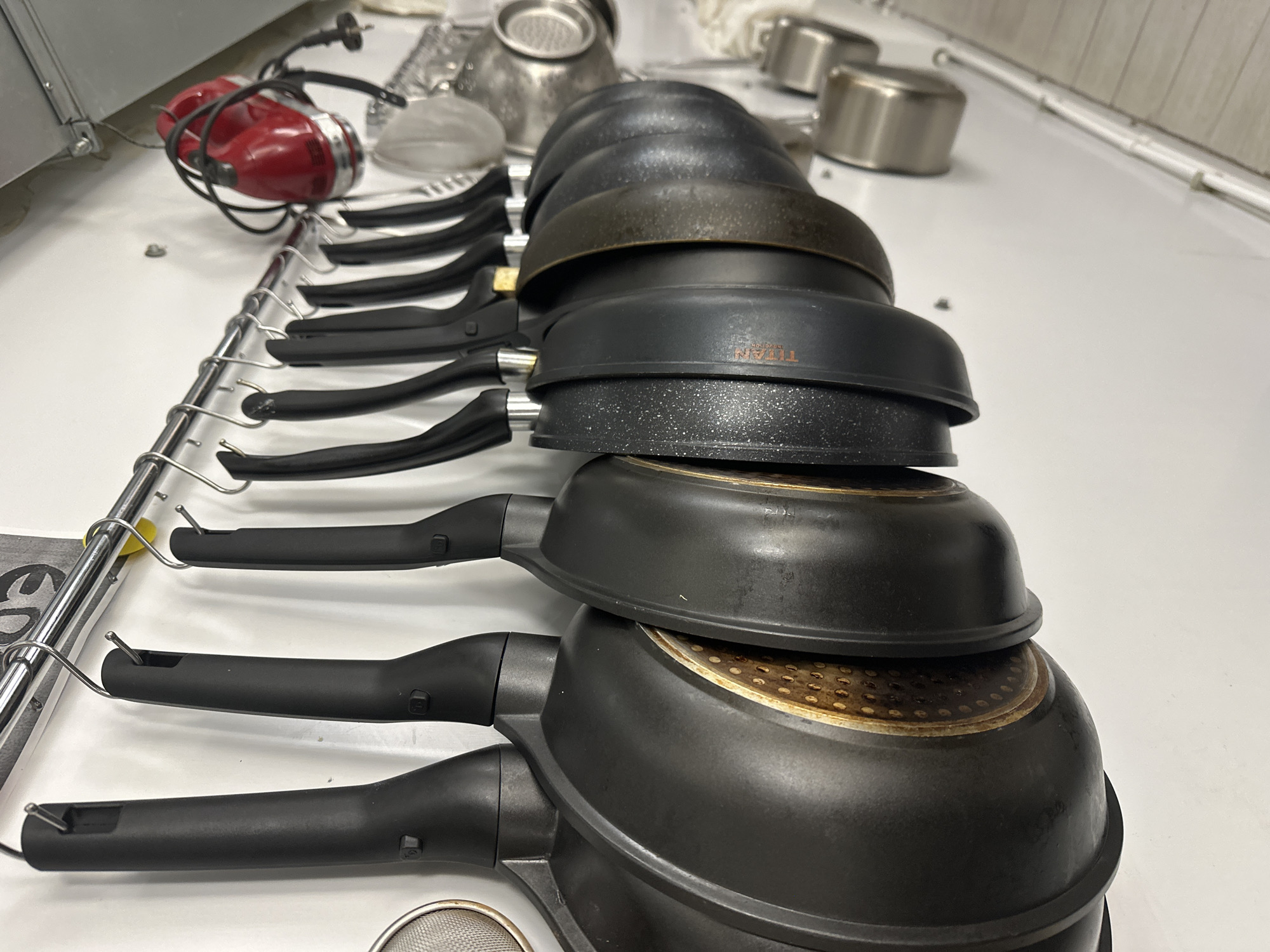 A row of black frying pans, stacked on top of each other, are positioned on a white surface. The pans have black handles and are hanging from a silver metal rack. A red hand mixer and various metal cooking utensils, including small pots and a strainer, are also present on the surface. The location is Новоглаголево, Russia.