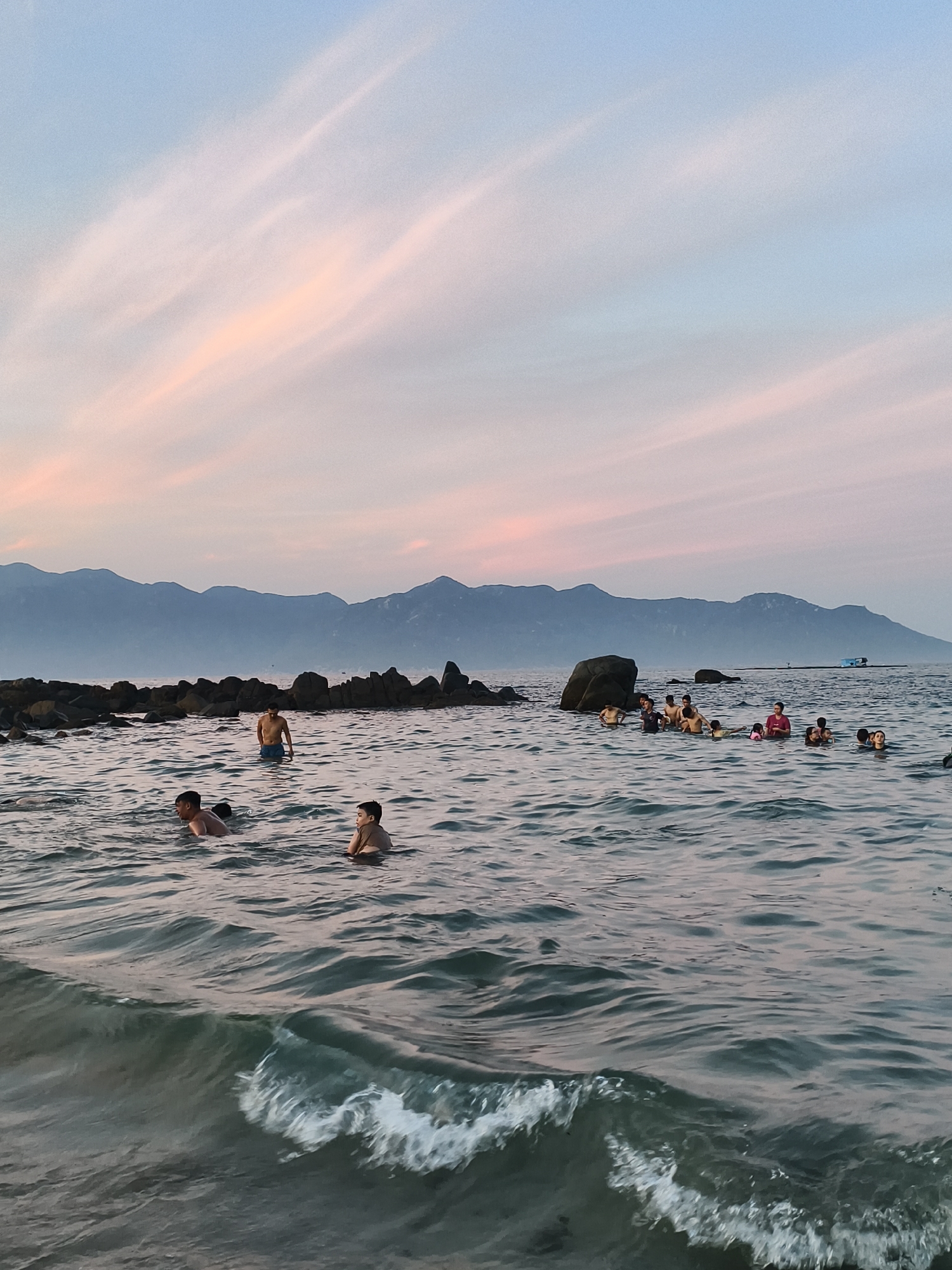 In Vĩnh Lương, Vietnam, at dusk, a group of people are swimming in the ocean near a rocky shoreline. The sky is painted with soft pink and blue hues from the setting sun, with wispy clouds stretching across it. The water is calm, with gentle waves lapping at the shore. In the foreground, a small wave is breaking, showing its foamy crest. Several individuals, including adults and children, are visible in the water, some swimming and others standing. In the background, a range of mountains forms a silhouette against the horizon, and a small blue boat is visible in the distance. The rocky formations along the coast are dark and weathered, jutting out from the water.