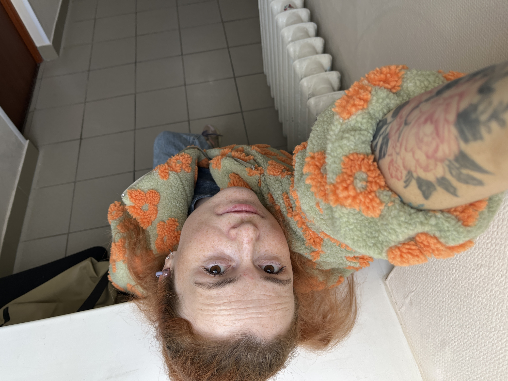 In an indoor setting, a person with fair skin and red hair, adorned with freckles and wearing a green fuzzy jacket with orange floral patterns, poses for a selfie. The subject is looking directly at the camera, with their left arm visible, showcasing a floral tattoo. The background consists of tiled flooring and a white radiator. The scene suggests a residential or public indoor space.