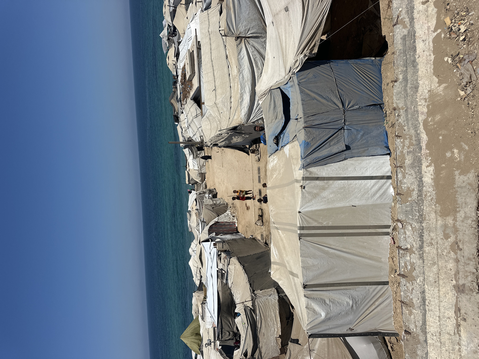 A high-angle shot captures a sprawling tent city situated on a sandy terrain adjacent to a bright blue ocean under a clear sky. Numerous temporary shelters, constructed from tarps and canvas in various shades of white, beige, and blue, dominate the scene. These tents are erected in a dense, irregular pattern, forming narrow pathways between them.
In the middle ground, three children are visible, two standing and one possibly sitting or lying down, on a sandy path. Further back, a man's face peers out from the entrance of one of the tents. A woman in dark clothing walks in the distance on the left. The environment is arid and dusty, with scattered debris visible on the ground. The ocean's edge is marked by a rough, uneven shoreline. The overall impression is one of a temporary, makeshift settlement.