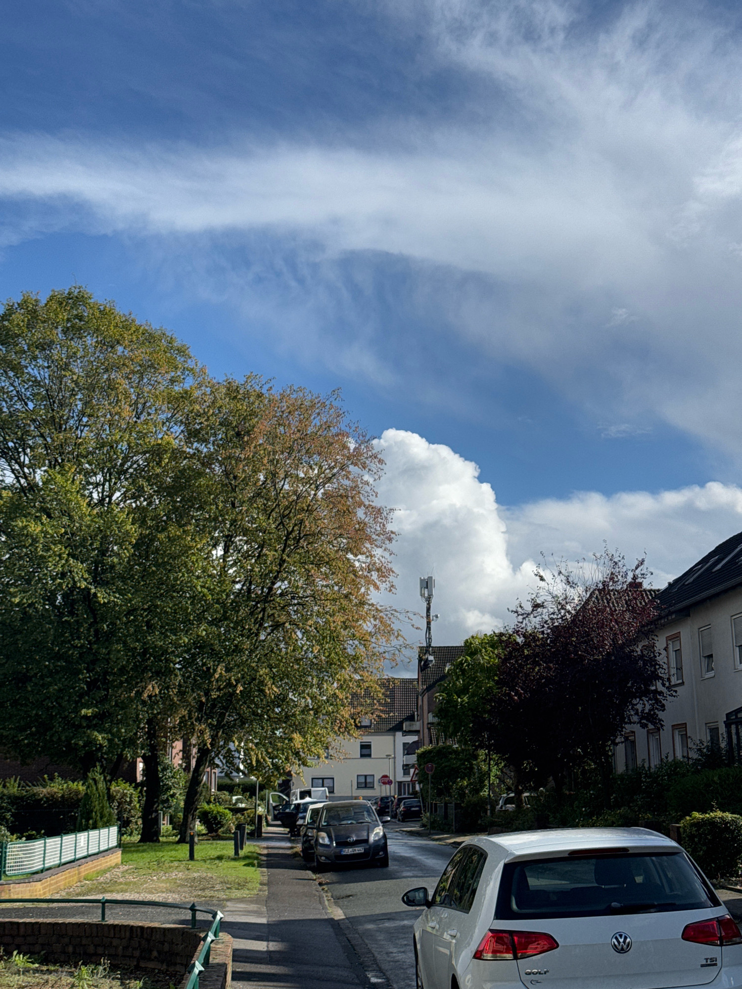 The image depicts a street scene in Cleves, Germany, under a partly cloudy sky. The foreground features the rear of a white Volkswagen Golf TSI. The street is lined with parked cars, including a dark-colored hatchback and a white van. On the left side of the street, there is a sidewalk bordering a grassy area with trees. A white fence runs along the edge of the grassy area. Buildings with white facades and dark roofs are visible on the right side of the street. A cell tower stands tall amongst the trees in the middle ground. The overall impression is a quiet residential street during the day.