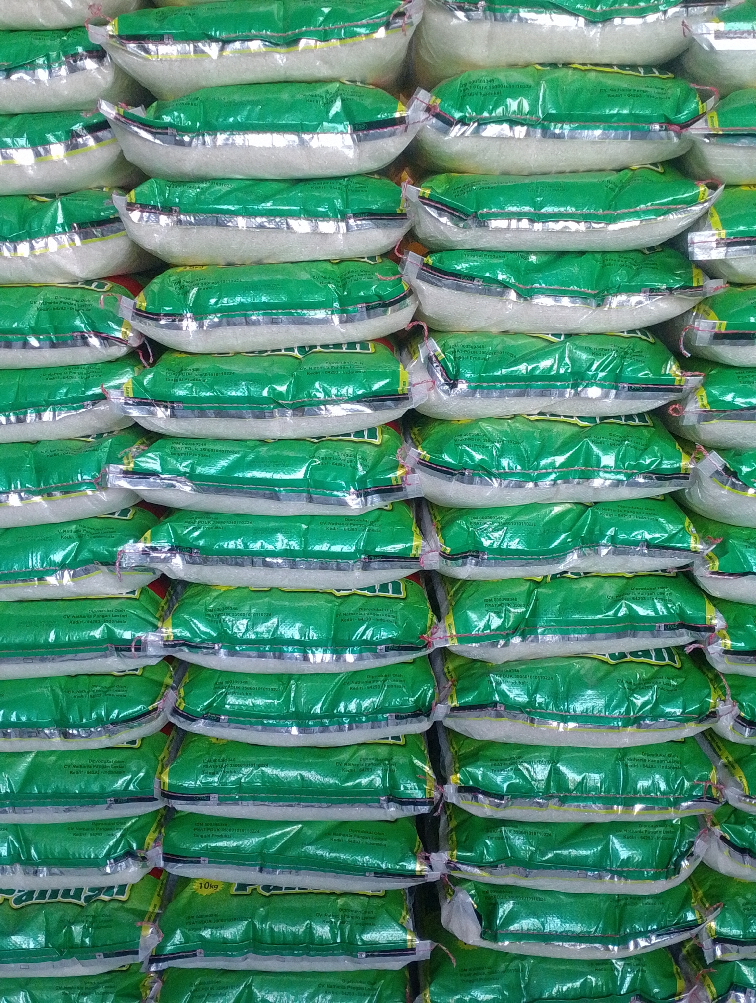 The image shows a tightly packed stack of rice bags, predominantly green with silver accents, in what appears to be a storage or retail setting. The rice bags are semi-transparent, revealing the white grains inside. Several bags display text in Indonesian, including "Diproduksi Oleh CV. Nathania Pangan Lestari" and "Kediri - 64283 - Indonesia." One visible bag indicates "10kg." The arrangement suggests a large quantity of rice is being stored or displayed. There are no people visible in the image, and the environment is focused solely on the stacked rice bags. The location is identified as Tungkaran Pangeran, Indonesia.