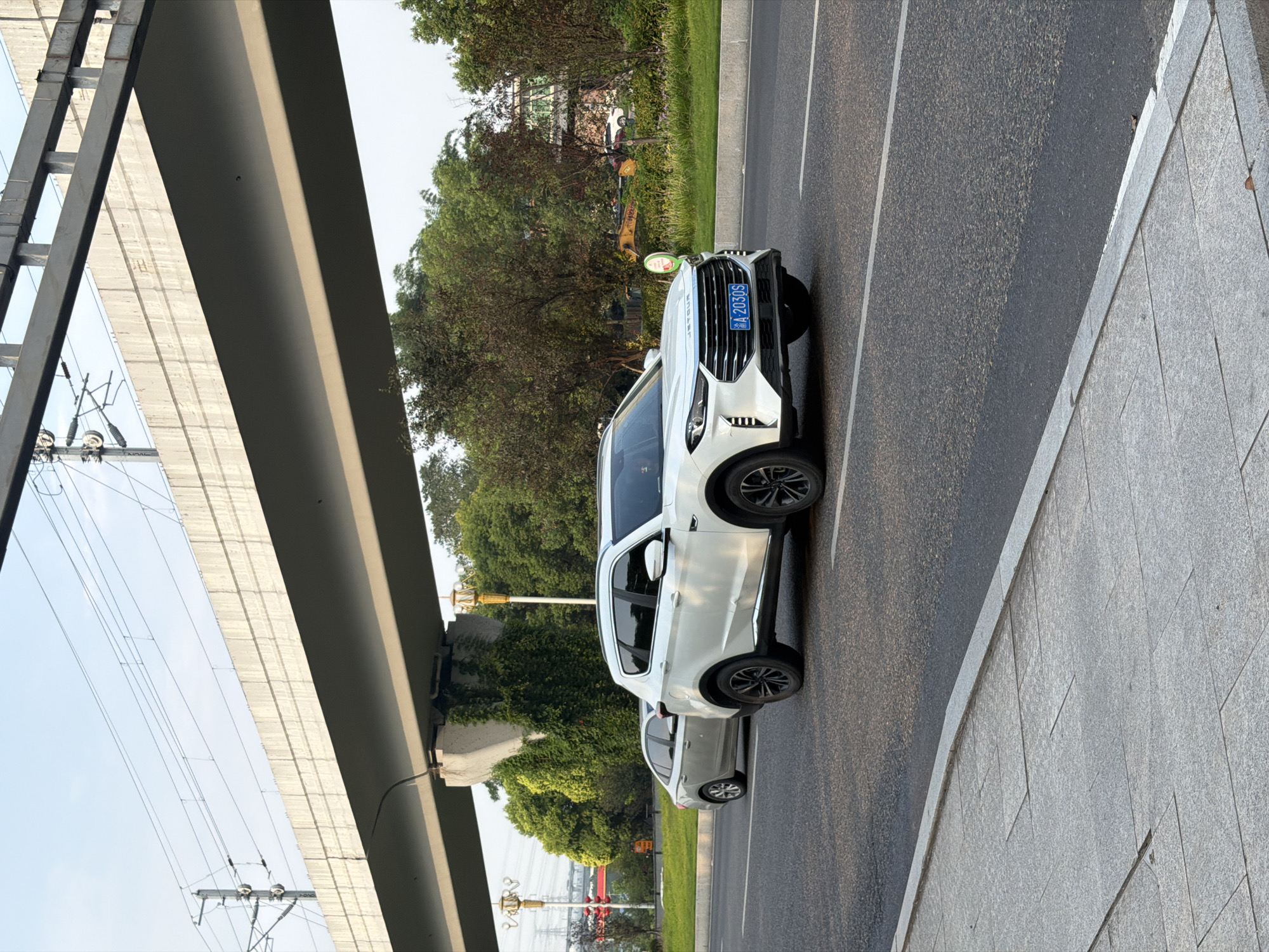 A white SUV is driving on a road in Jiangjin, China, under a concrete overpass with power lines. The SUV has a blue license plate that reads "A 2030S". A silver sedan is driving behind the SUV. The road is paved and has white lane markings. A sidewalk made of granite pavers is to the left of the road. Trees and vegetation are visible on the right side of the road, with buildings and industrial structures in the background. The sky is clear and blue.