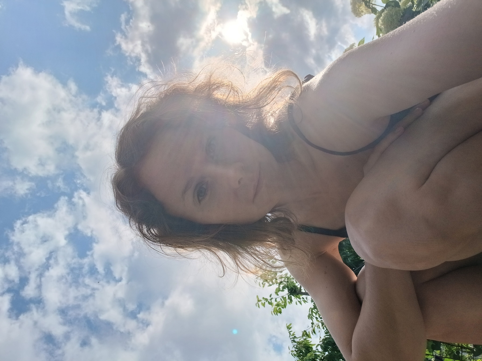 In Maslova Pristan', Russia, a woman with auburn hair is captured in a low-angle selfie outdoors. She is crouching, with her arms and legs pulled in towards her body. The woman is wearing a dark-colored top, possibly a bikini top. Her hair is windswept, and the sunlight is glinting off her face and hair, creating a lens flare effect. Behind her, the sky is filled with a mix of blue sky and white clouds. Lush green foliage, including trees and some white flowers, is visible to the left and right of the woman, suggesting she is in a garden or park setting. The overall atmosphere is bright and natural, with the sun being a prominent element in the background.
