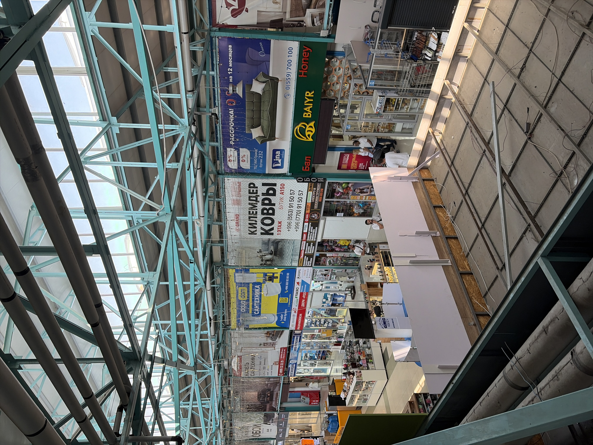 The image depicts the interior of a large, open-plan marketplace in Bishkek, Kyrgyzstan. The structure features a high, translucent ceiling supported by a teal-colored metal frame, allowing natural light to illuminate the space. Various stalls and shops are visible on the ground floor, displaying a diverse range of goods, including furniture, home goods, and electronics, indicated by signage in Cyrillic script. Several people are seen browsing or working in the marketplace. The upper levels of the market are also visible, with walkways and additional stalls. The overall impression is of a bustling commercial environment.