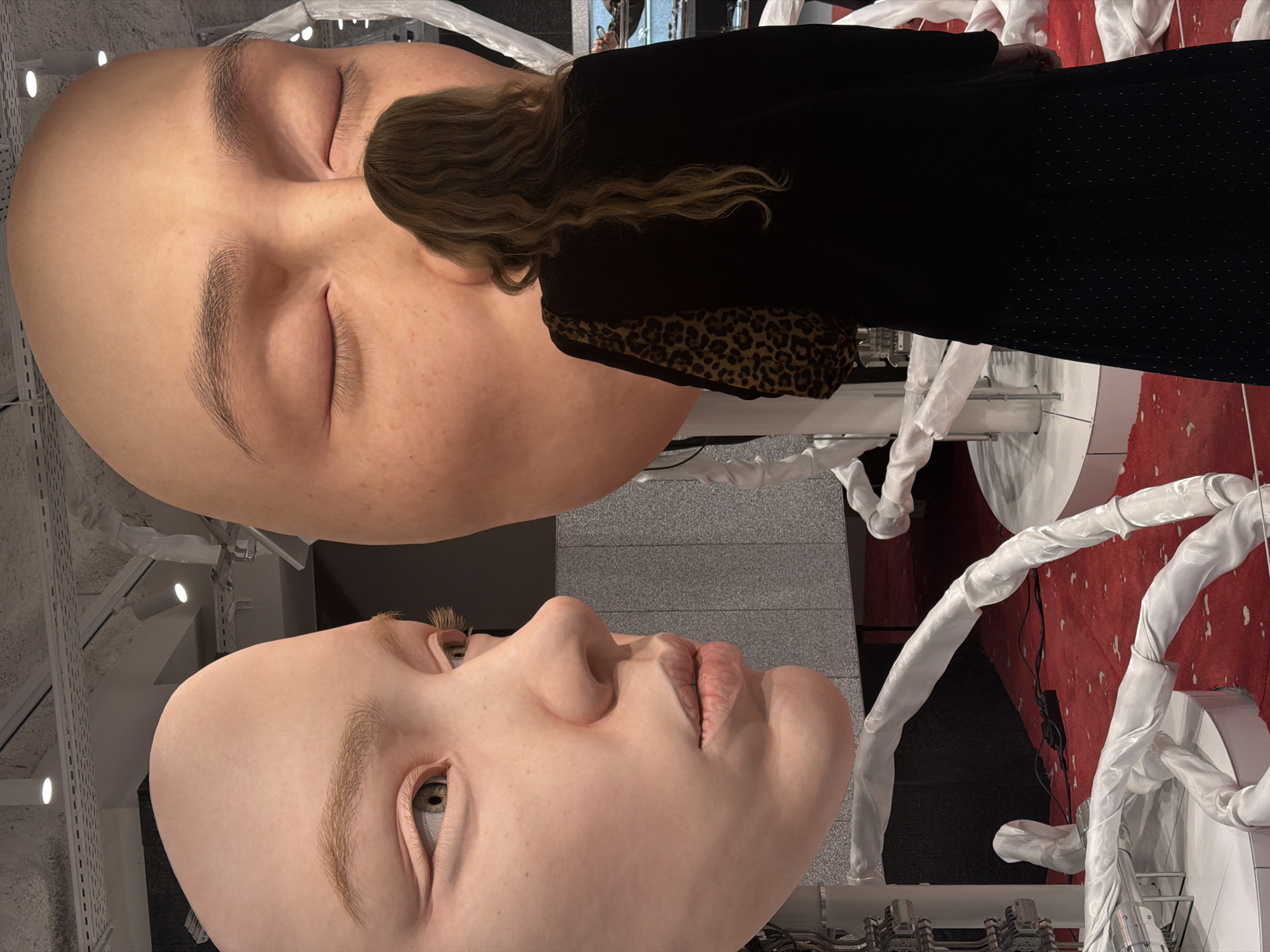 The image captures a surreal scene at an art installation, likely in Paris, France. Two oversized, hyperrealistic human heads dominate the view. The upper head is in repose, with closed eyes and a serene expression. A woman stands near it, her dark dress and leopard-print trim visible, with her wavy brown hair falling near the head's face. The lower head, with open eyes, gazes upward, its features intricately detailed. The installation features white fabric-covered structures resembling stylized limbs or pipes, adding to the dreamlike quality. The environment appears to be an art gallery or exhibition space, with red and grey surfaces, and some technical equipment visible.
