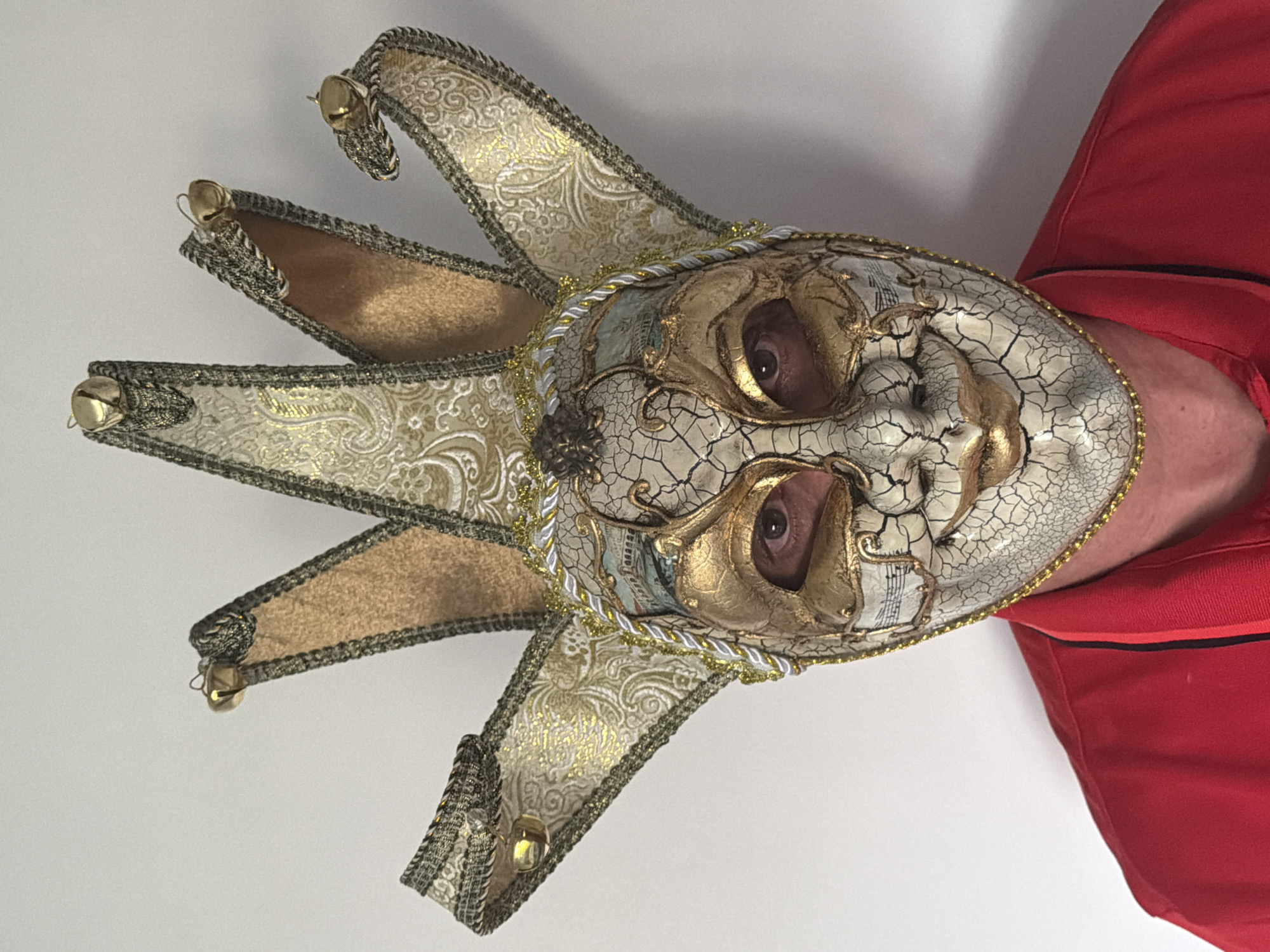 The image shows a person wearing a jester mask and a red shirt, set against a plain white background. The mask is ornate, featuring a gold and white patterned crown with small bells. The mask itself is white with gold accents and has a cracked texture. The person's eyes are visible through the eyeholes of the mask. There are no discernible environmental cues or any indication of the specific location in Esch-sur-Alzette, Luxembourg. The scene is static and focuses solely on the individual and their attire.