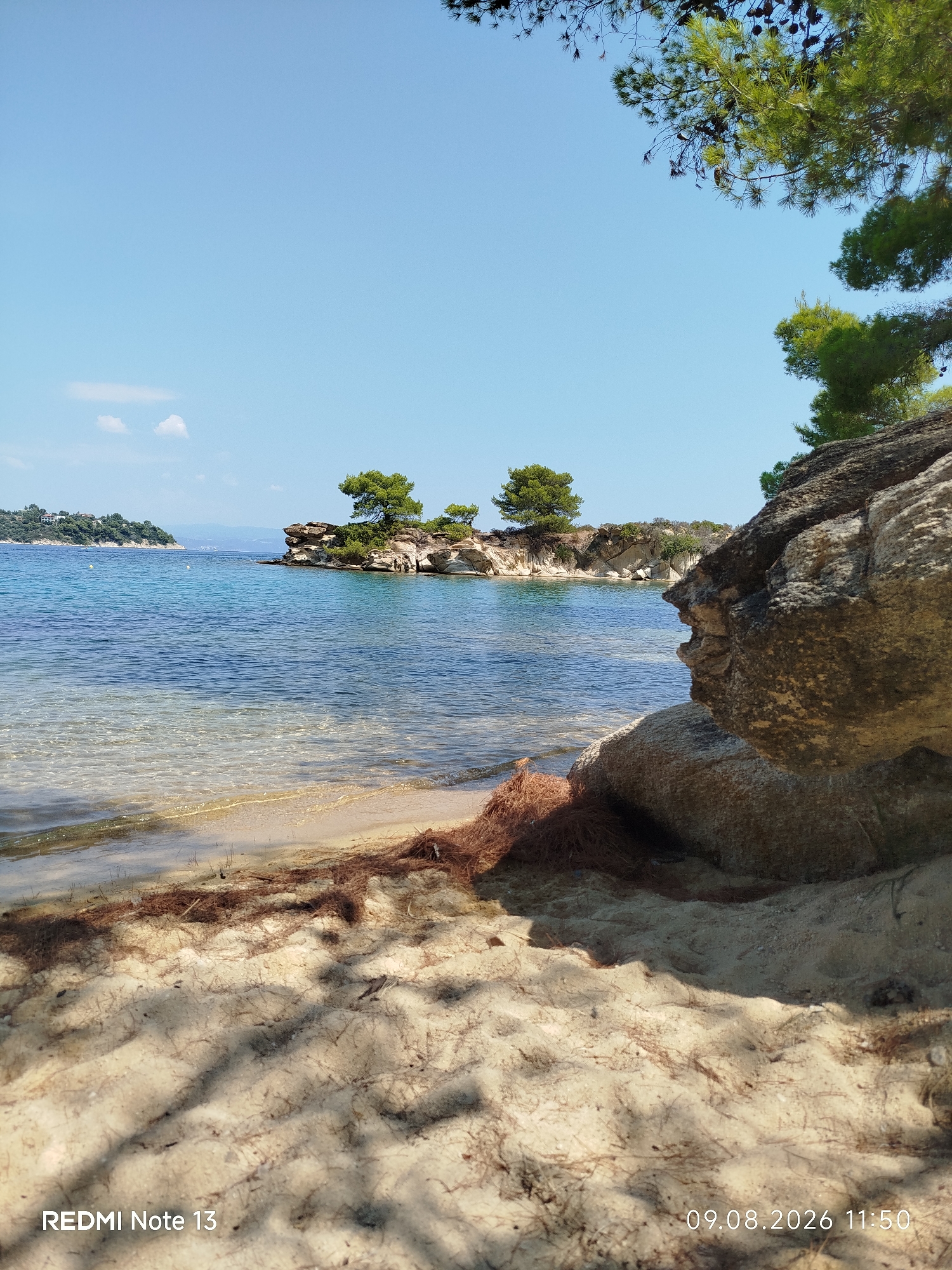 A sunny day at a sandy beach in Sithonia Municipal Unit, Greece. The foreground is dominated by sand with scattered pine needles and shadows cast by unseen foliage. A large, textured rock formation occupies the right side of the frame. The crystal-clear turquoise water laps gently onto the shore, revealing the sandy bottom. In the middle ground, a small rocky island with sparse green trees stands against the calm sea. Further in the distance, a landmass with buildings and trees is visible on the horizon under a clear blue sky with a few wispy clouds. The timestamp "09.08.2026 11:50" and the text "REDMI Note 13" are present in the bottom left and right corners, respectively.