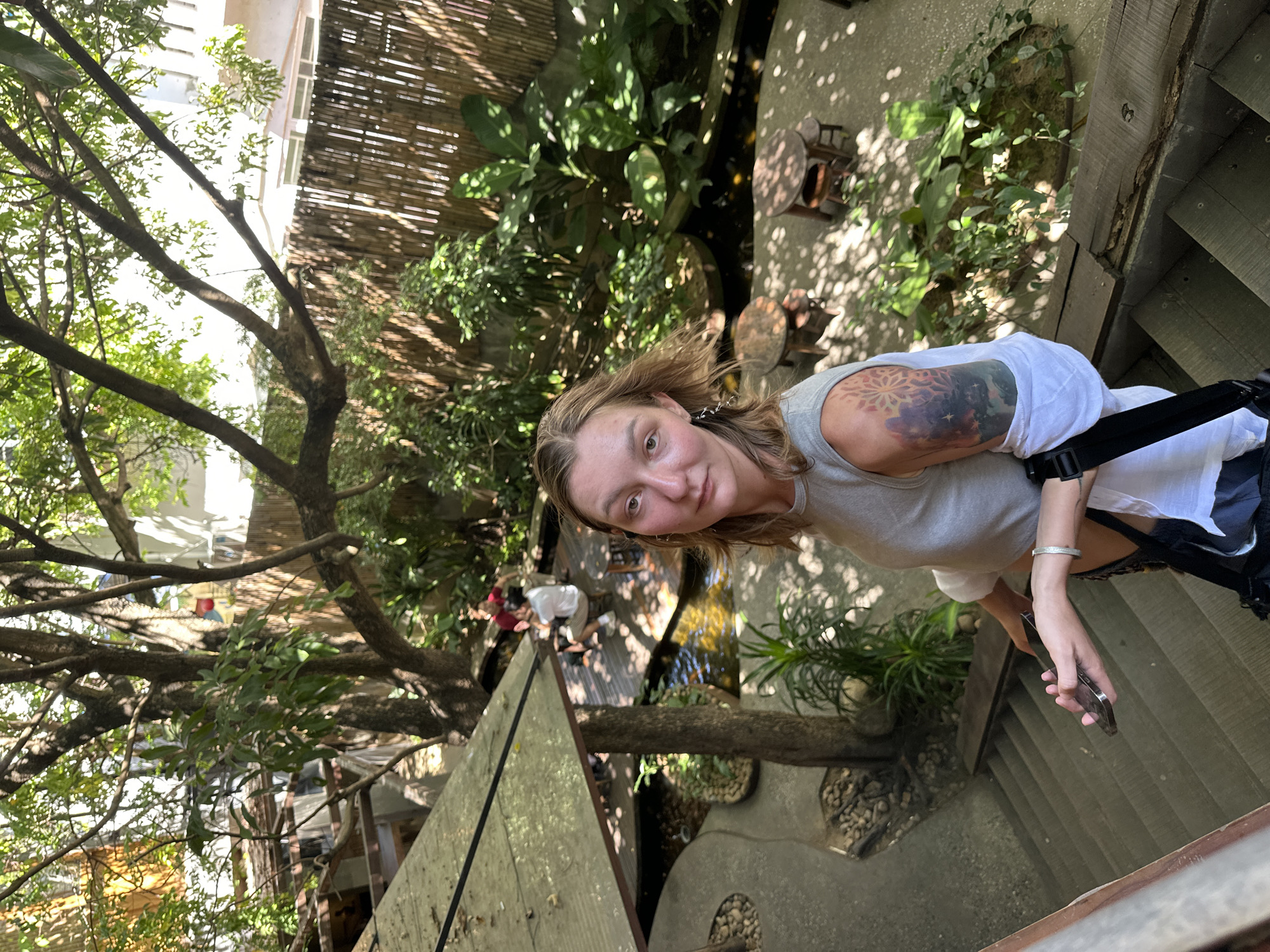 In Đà Nẵng, Vietnam, a woman with blonde hair and a tattoo on her left arm stands on a stairway, looking at the camera. She is wearing a gray tank top and a white shirt. In the background, there is a lush garden with trees, plants, and a pond. Several small wooden tables and chairs are scattered throughout the garden. Two people are seated at a table by the pond. The scene appears to be a tranquil outdoor cafe or restaurant.