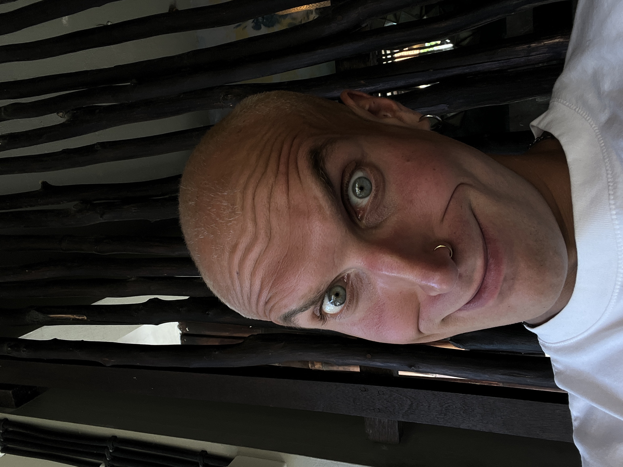 A close-up, slightly distorted selfie is shown, taken outdoors in a dimly lit setting. The subject is a bald individual with light-colored hair, a light complexion, and fair skin with visible pink undertones on the cheeks. They have striking blue eyes, a wide, unnerving grin, and wrinkles on their forehead. A small gold hoop is visible in their left eyebrow, and a silver hoop earring is in their left ear. The background consists of dark, vertical wooden poles, creating a sense of enclosure and depth. The overall impression is one of a playful or exaggerated facial expression against a rustic backdrop. The scene is likely outdoors in the city of Mengwi, Indonesia, given the ambient lighting and natural elements.