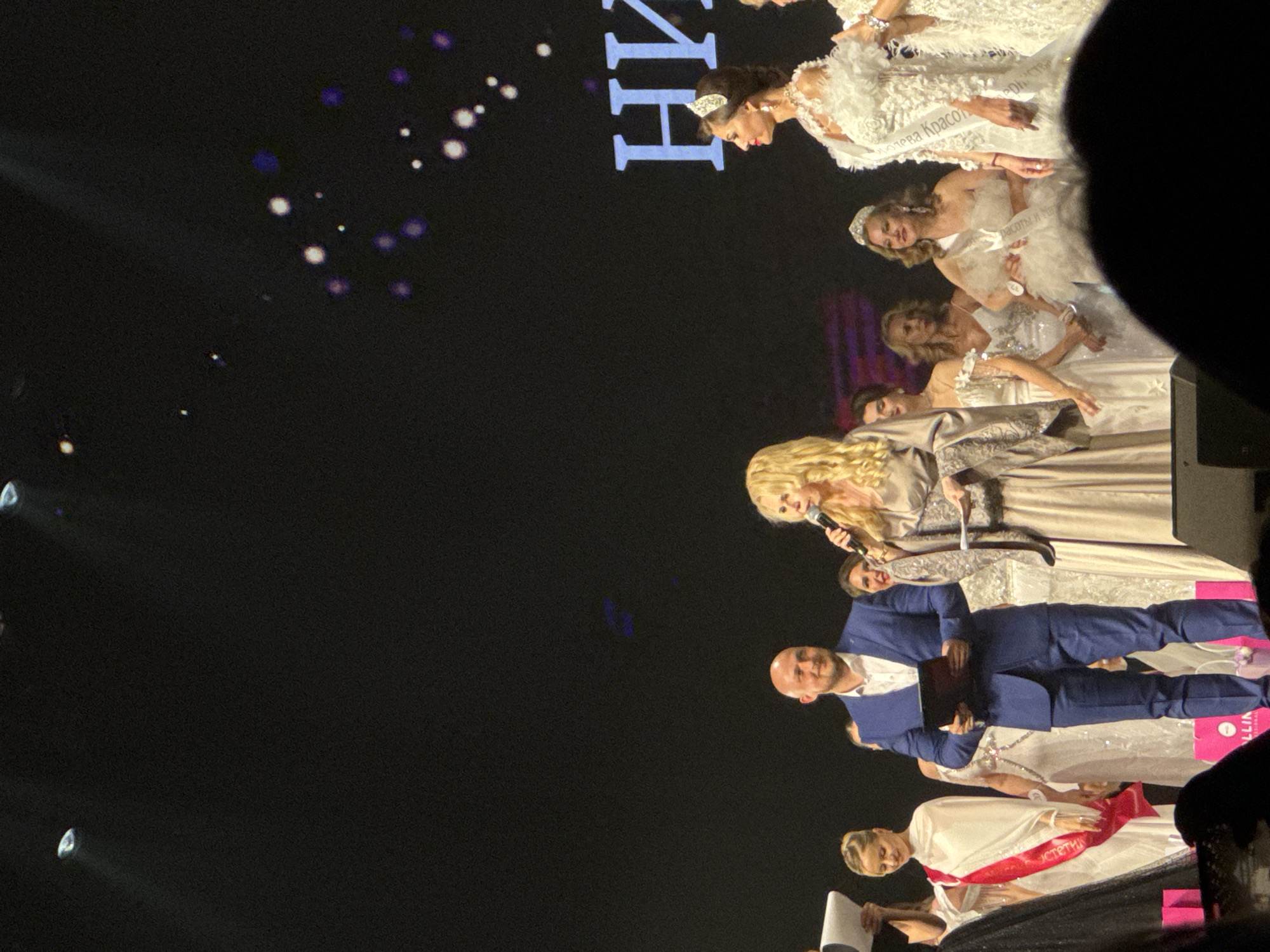 Here is a detailed description of the image:
The image shows a stage in an entertainment venue, likely in Unknown, Unknown, with a group of people, mostly women, dressed in formal wear. Some of the women are wearing white gowns and sashes with text in Cyrillic. A blonde woman stands at the front with a microphone, seemingly speaking or singing. A man in a blue suit stands to her side holding a tablet or notebook.
The background is mostly dark with some light spots and Cyrillic text at the top. There are stage lights visible, adding to the ambience. The overall impression is that of a beauty pageant or a similar formal event taking place on a stage.