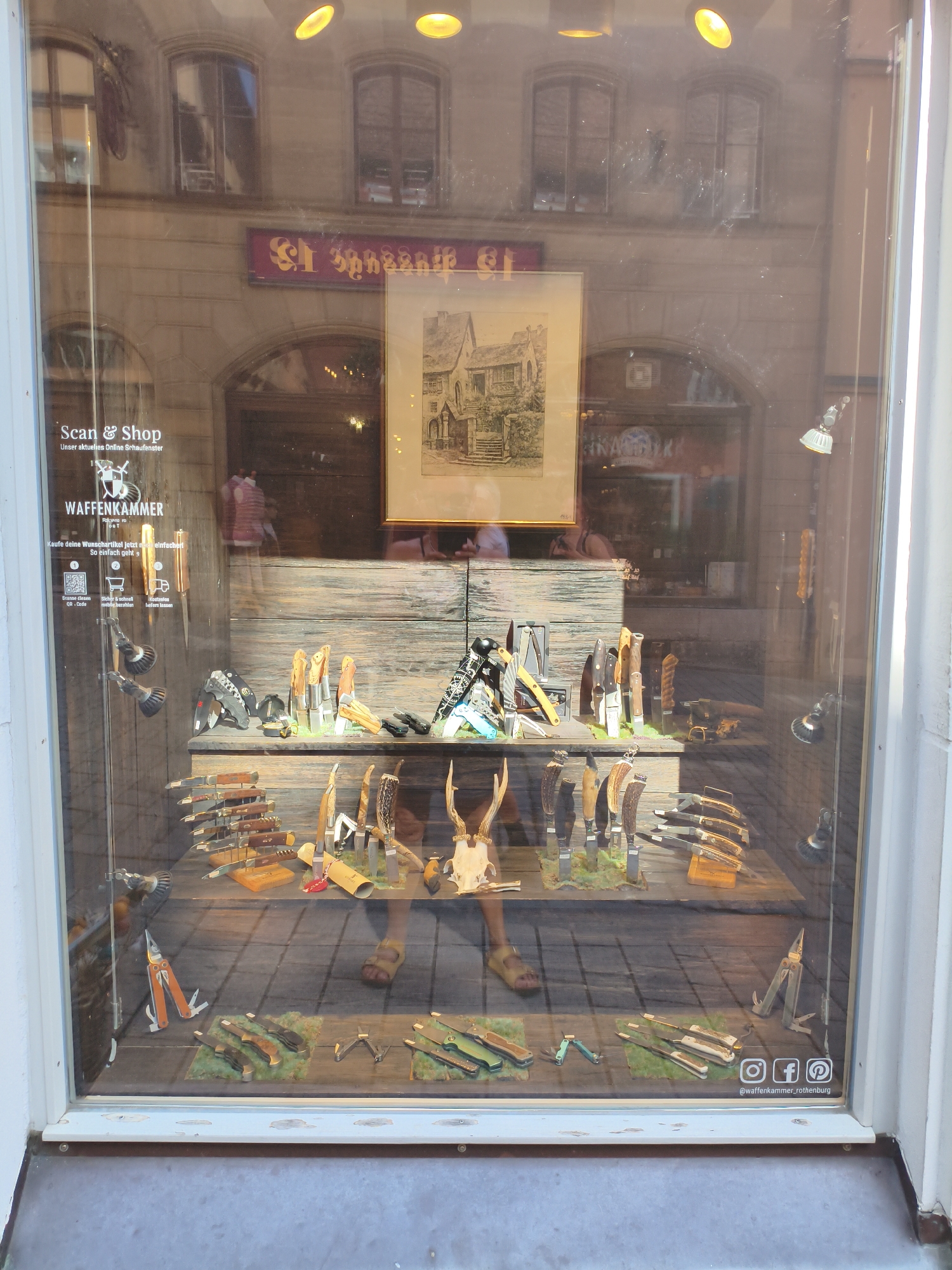 This image shows a street-level window display of a shop in Rothenburg ob der Tauber, Germany. The shop appears to be a knife store, as evidenced by the variety of knives and related items on display. The display is set up on wooden shelves and includes knives with various blade shapes and handle materials, as well as multi-tools and what appear to be deer antlers. A framed drawing of a building is also visible in the upper portion of the window. The text "Scan & Shop" and "WAFFENKAMMER" are visible on the window, indicating the shop's name and a QR code for online shopping. Social media icons for Instagram, Facebook, and Pinterest are present along with the shop's handle "@waffenkammer_rothenburg". The reflection in the window shows buildings and lights from across the street. A person's legs and feet are visible wearing sandals, suggesting they are the shopkeeper or a customer standing inside or just in front of the display.