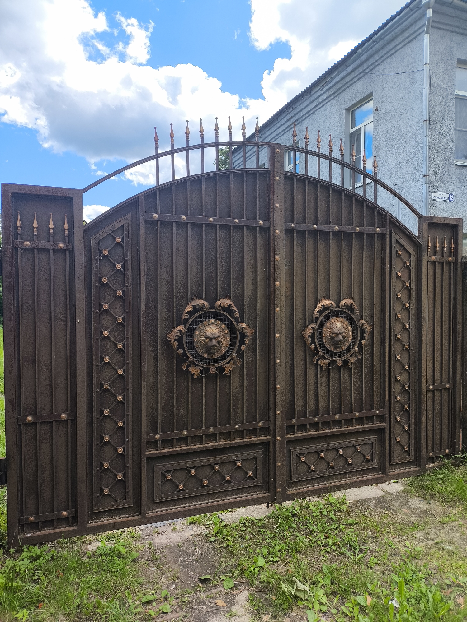 This is a vertical, medium-angle, outdoor, eye-level shot of a dark brown wrought iron gate in Taldom, Russia. The gate has two arched main panels, each decorated with a circular medallion featuring a lion's head. The main panels are flanked by narrower vertical gate sections, also made of wrought iron, and topped with pointed finials. The gate is set against a building with a light gray, textured facade and a dark roof. A street sign with Cyrillic lettering and the number "12" is visible on the building's wall. The ground in front of the gate is covered in grass and a concrete path. The sky is blue with scattered white clouds.
