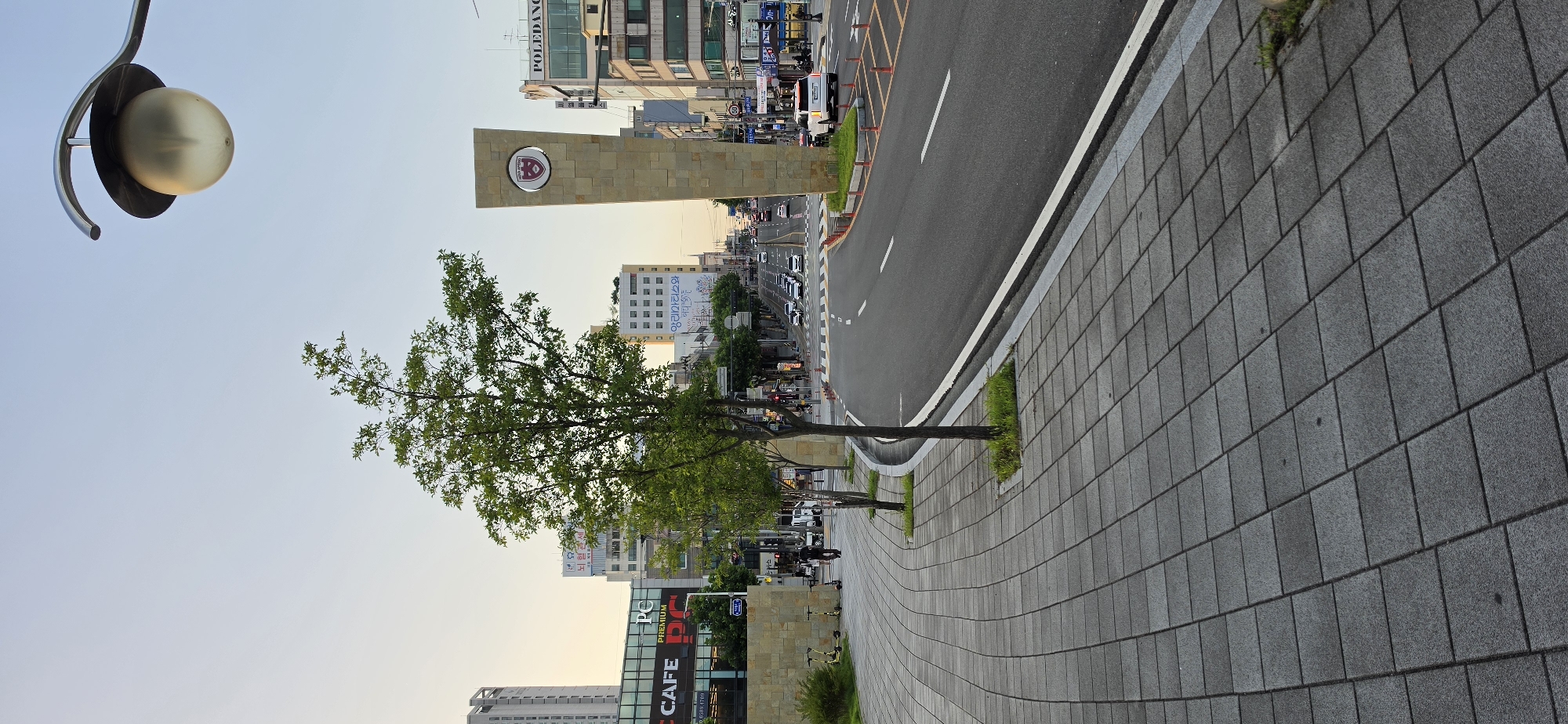 This is a street-level, eye-level, medium-shot photograph of a street in Cheongju-si, South Korea, likely taken in the late afternoon given the soft, warm light. The foreground features a gray paved sidewalk with a repeating square tile pattern that curves to the right. A young tree with green foliage is planted in a small grass patch on the edge of the sidewalk. To the right of the sidewalk is a paved road with white lane markings.
In the midground, a large, stone monument shaped like a tall, rectangular pillar stands prominently. It has a circular emblem at its top with a red and white crest. Across the street, traffic is visible, with several cars driving in both directions. Buildings line the street on either side, including a multi-story building with "POLEDANG" written on it. A prominent building with a large "CAFE PC" sign is visible on the left.
In the background, more buildings and streetlights are visible, extending down the street towards the horizon. The sky is a clear, pale blue with a hint of yellow near the horizon, suggesting either sunrise or sunset. A streetlamp with a globe-shaped light fixture is positioned in the upper right corner of the frame. A person is walking on the sidewalk in the midground, and several electric scooters are parked near the stone wall on the left.