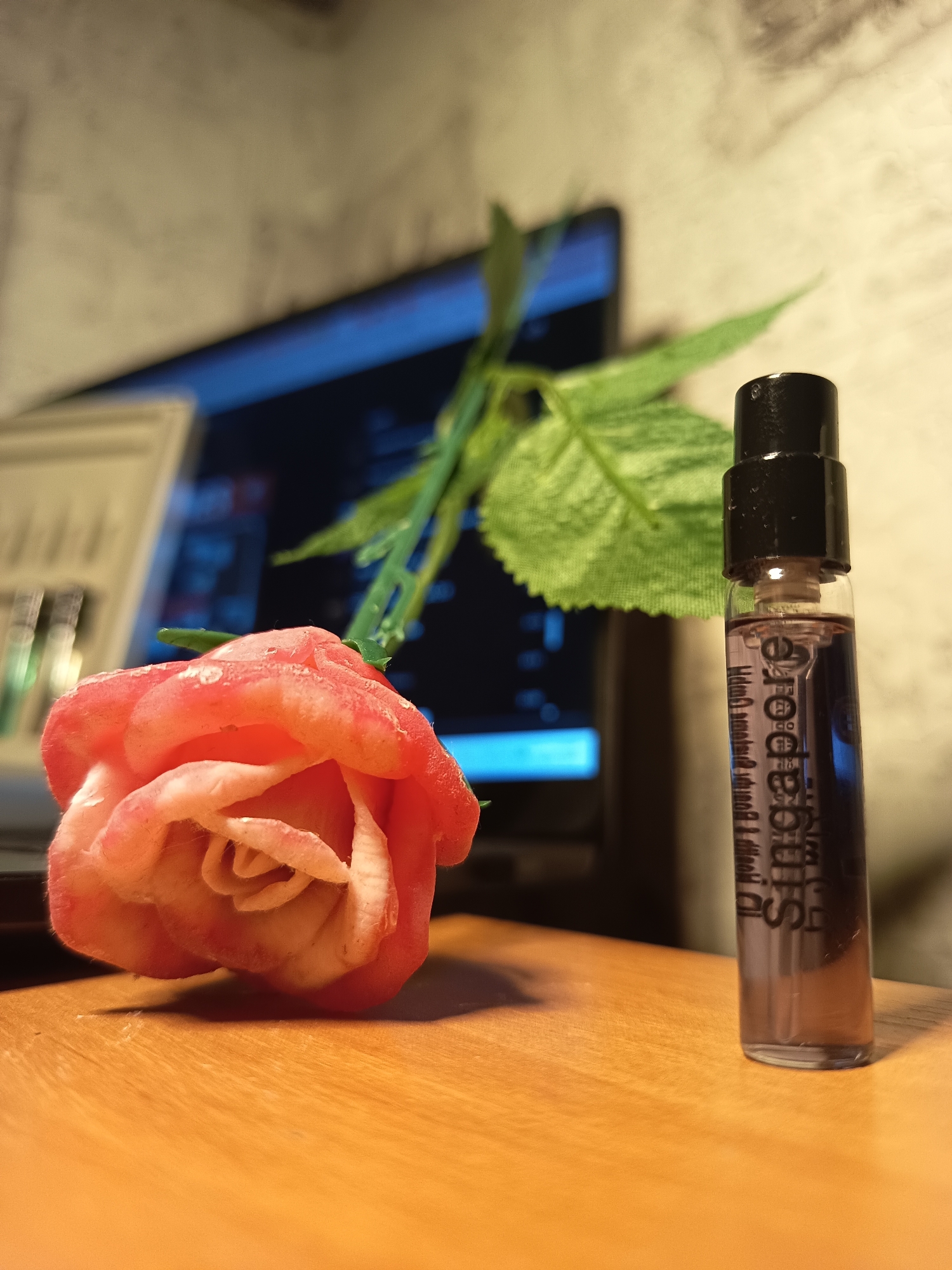 A close-up, eye-level shot shows a single pink and white artificial rose placed on a wooden surface. To the right of the rose, a small, clear glass perfume sample bottle with a black cap stands upright. The bottle contains a dark purple liquid. Text is visible on the bottle, appearing to be a brand name or fragrance description, though it is partially obscured and difficult to read clearly. In the background, out of focus, is a laptop screen displaying what appears to be a grid-like interface or data, and a blurred green leaf from a plant. The overall lighting suggests an indoor setting, possibly a desk or table, with a textured wall in the background. There are no people or animals present in the image. The context of Izmail, Ukraine, is not visually represented in the provided image.