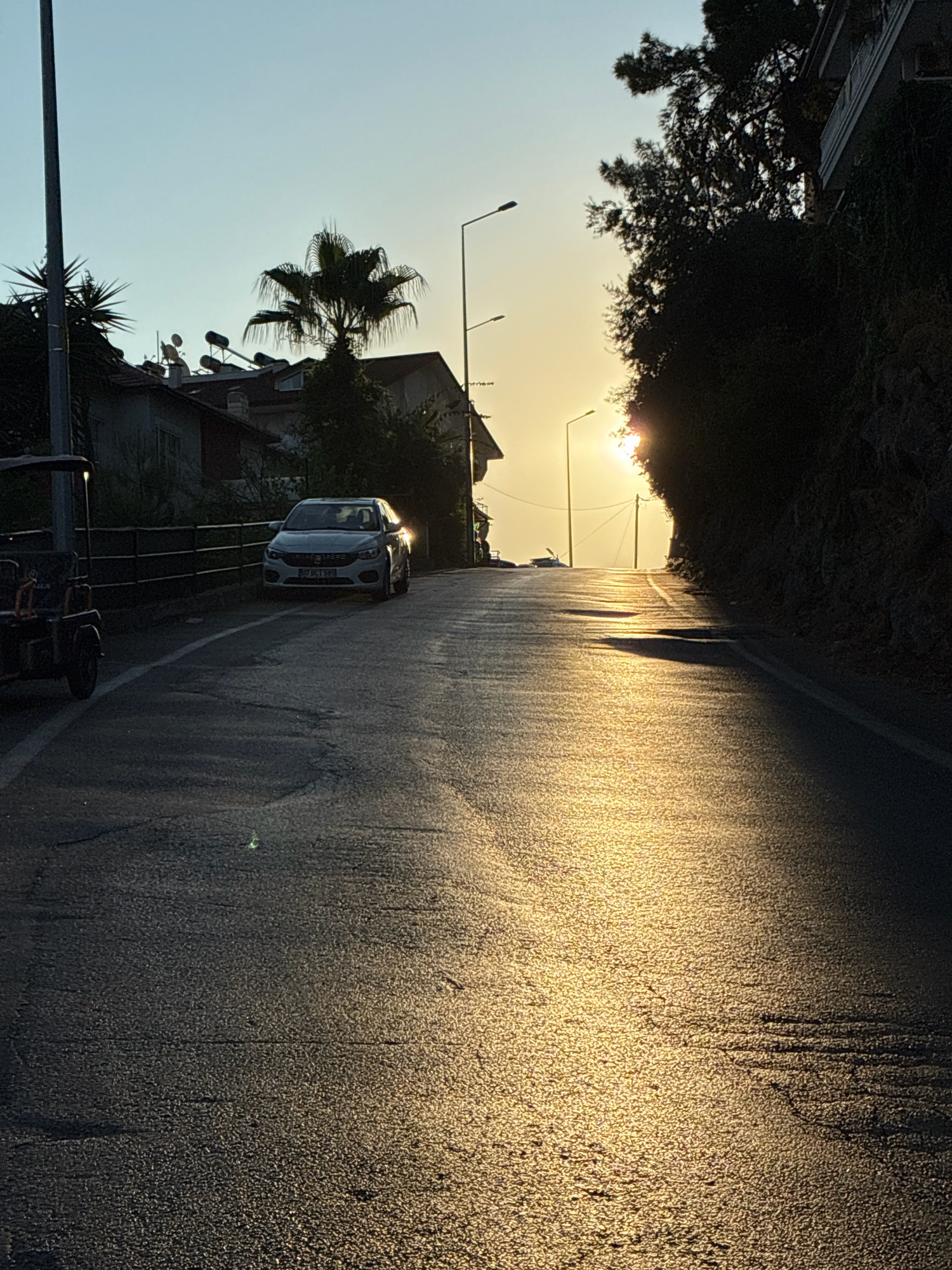 The image captures a sun-drenched road winding uphill in Alanya, Turkey, likely during the golden hour. The road surface is asphalt, reflecting the low-angled sunlight, creating a shimmering effect with visible cracks and texture. On the left side of the road, a white sedan is parked, its headlights illuminated. Further up the left side, a small, open-air vehicle with a canopy is partially visible. Beyond the vehicles, residential buildings with satellite dishes and solar water heaters are nestled amongst trees, including a prominent palm tree. Streetlights are positioned along the road. The right side of the road is bordered by a steep, tree-covered embankment, with a building partially visible at the top. The sky is a clear, pale blue, transitioning to a warm yellow hue near the horizon where the sun is setting, peeking through the trees. The overall scene depicts a quiet street in a Mediterranean coastal town.