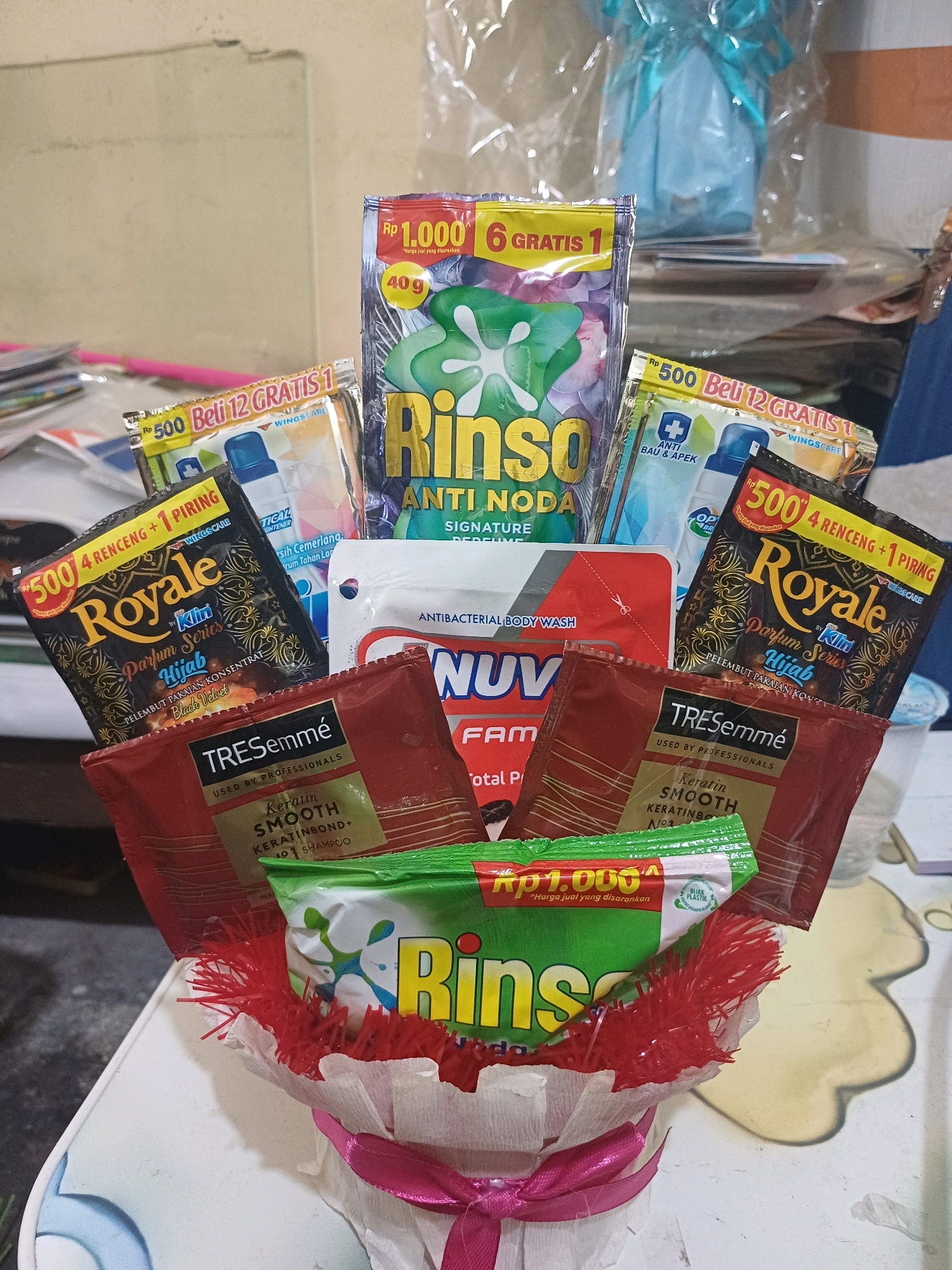 This is a close-up, eye-level, indoor photograph of a gift arrangement, likely a "snack bouquet" or "product bouquet." The arrangement is composed of various personal care and cleaning products, predominantly sachets and small packages.
Dominating the center is a large sachet of Rinso laundry detergent with a "signature perfume" variant, featuring green and purple graphics and the brand name in large yellow lettering. Surrounding it are several smaller sachets.
On the left, two black and gold sachets of "Royale Parfum Series Hijab Black Velvet" are visible. To their right, there's a sachet of "NUU FAM" antibacterial body wash. Below the Rinso and to the right of the Royale sachets, a red sachet of "TRESemmé Keratin Smooth Shampoo" is prominent. In front of the TRESemmé sachet, another green Rinso sachet is visible. Other smaller, less distinct sachets are also present, including what appears to be a fabric softener.
The products are arranged in a cylindrical container, partially obscured by red and white tissue paper and a pink ribbon tied in a bow at the bottom. The background is blurred, showing a light-colored wall and some plastic wrapping, possibly from another gift.
The image does not provide any specific visual cues that would definitively place it in Dumai City, Indonesia. The products themselves, such as Rinso and Royale, are widely available in Indonesia, but there are no local landmarks, signs, or identifiable features that indicate a specific city. The environment appears to be a domestic or retail setting.