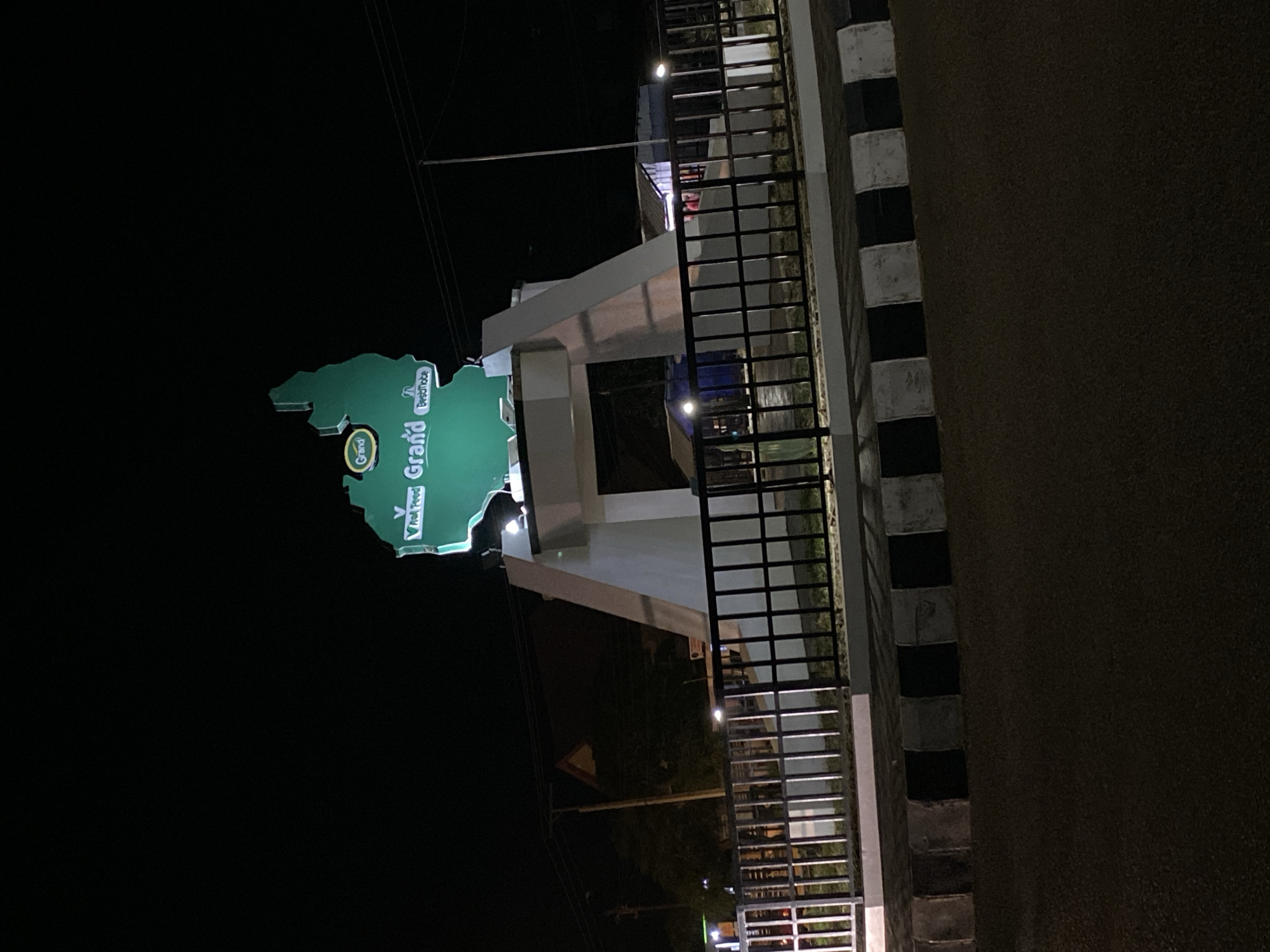 A monument shaped like a map of Nigeria is illuminated at night in Bukuru, Nigeria. The monument is green and features the words "Grand" and "BestNobe" with a logo above. It stands on a white concrete base with black and white striped borders around it. A black metal fence encloses the monument. In the background, there are buildings with lights, streetlights, and power lines. The scene is captured from a low angle, looking up at the monument.