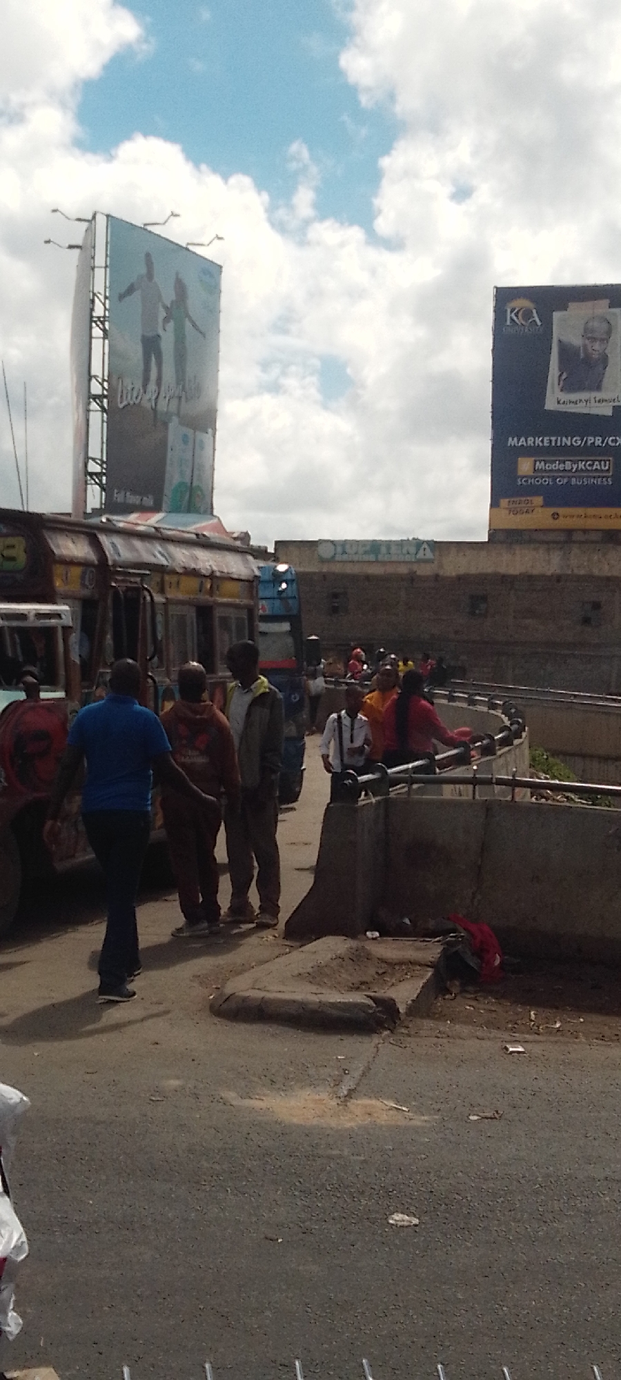 The image captures a detailed urban scene in Nairobi, Kenya, specifically identifiable by the "KCA University" billboard, likely situating the area near the university campus on Thika Road. Under a partly cloudy sky with visible blue patches, two large billboards dominate the upper frame; the left one displays two silhouetted figures with outstretched arms and text "Lite up your life," while the right billboard advertises "KCA University School of Business" featuring an image of "Kaimenyi Samuel." In the foreground, on an asphalt road surface littered with debris, approximately ten individuals are present. A man in a blue polo shirt and dark trousers walks towards the left. To his right, two other men stand, one in a reddish-brown hoodie and another in a light-colored jacket. Multiple "matatu" public transport vehicles, including a multi-colored one, are partially visible on the left. A curved concrete pedestrian overpass or barrier, topped with a dark metal railing, spans the mid-ground, with several more pedestrians visible along it, including a person in a red top and another in a yellow garment. A low concrete barrier and a patch of exposed dirt are in the lower right foreground, where a red fabric item rests. Buildings are visible in the background, including one with "UP TEN" signage. The scene indicates a busy daytime thoroughfare with pedestrian and vehicular activity.