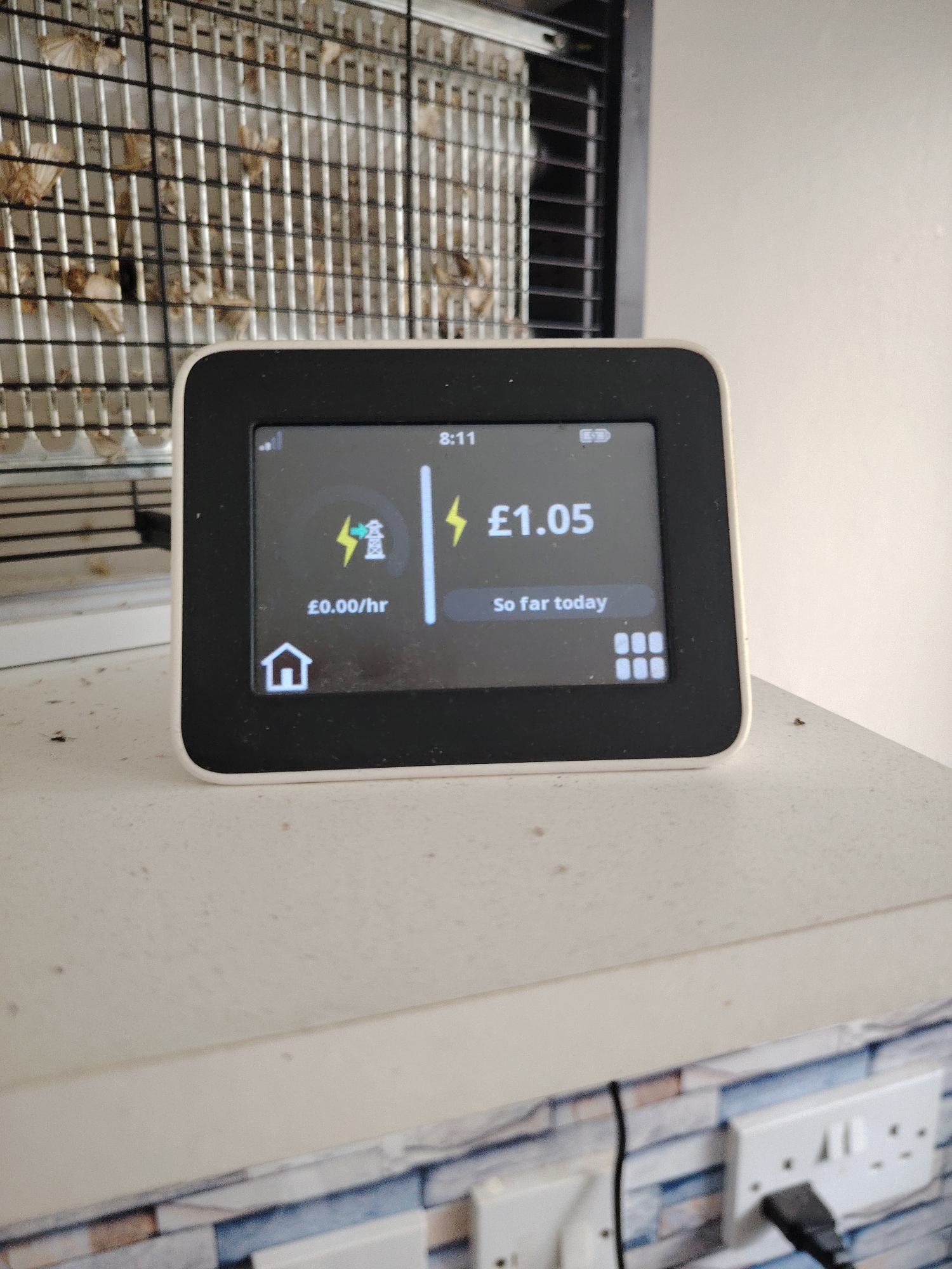 The image displays an indoor scene, likely within a domestic or utility area in Huntingdonshire, United Kingdom. Central to the frame is a white-bezeled smart energy monitor with a black digital screen, displaying the time as "8:11" in the upper right. The screen shows a current energy usage of "£0.00/hr" on the left, next to a green lightning bolt and a power pylon icon, indicating very low or no current consumption. On the right, a yellow lightning bolt precedes "£1.05" with the text "So far today," indicating the accumulated electricity cost for the current day. A home icon is visible in the bottom left corner, and a grid of six squares (menu icon) is in the bottom right.
Behind the energy monitor, positioned on a surface, is a rectangular insect zapper or trap, characterized by a fine metal grid. Numerous small, light-brown, desiccated organic forms, consistent with dead insects (likely moths), are visibly entangled within the grid. To the right of this device, a black, slatted structure is partially visible.
The energy monitor rests on a light beige or off-white countertop or shelf, which shows signs of dust and minor dark specks. Below this surface, a wall is visible, adorned with a decorative pattern resembling rectangular tiles in varying shades of blue, grey, and white. Two white UK-standard double electrical outlets are mounted on this wall. A black plug is inserted into the rightmost socket, with its cable extending downwards, presumably connecting to the energy monitor or another unseen device. The left socket appears empty.