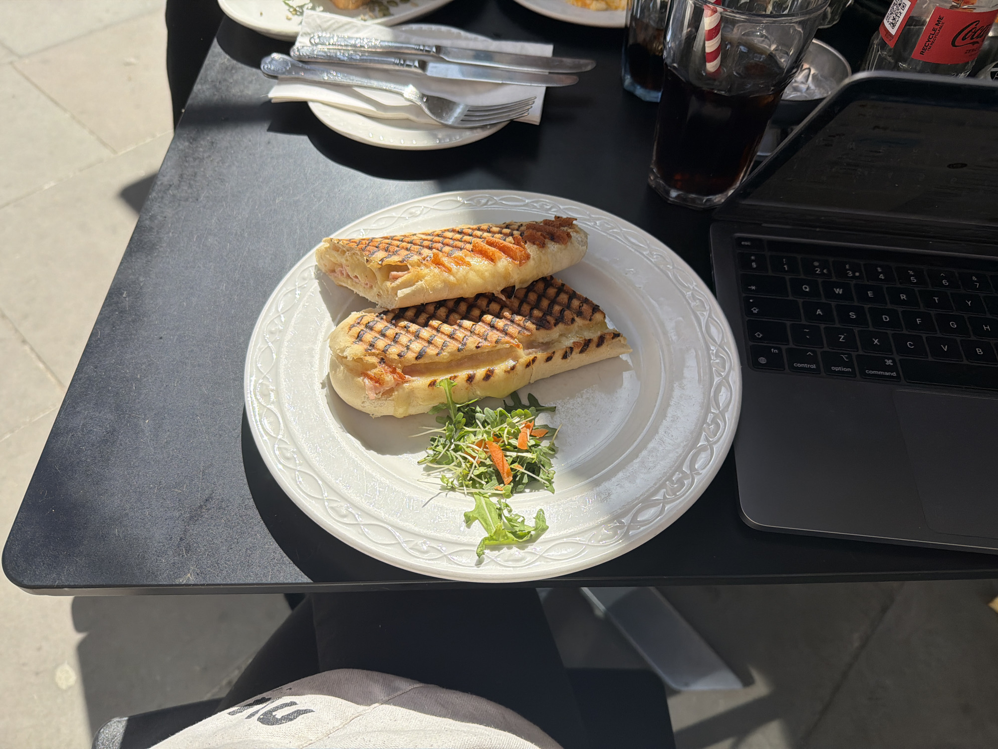 An overhead, slightly angled shot captures a casual outdoor dining and work setup on a sunny day in London. The scene is set on a black table, brightly illuminated by strong sunlight that casts distinct shadows.
In the foreground, a white plate with an ornate rim holds a grilled panini, cut into two halves, revealing a filling of ham and cheese. A small, fresh side salad of arugula, sprouts, and shredded carrots accompanies the sandwich. To the right of the plate, a black laptop (identifiable by the "command" key) is open, its keyboard showing keys like "esc," "control," "option," "command," along with letters (QWERTY, ASDFGH, ZXCVB) and numbers (1-7), including currency symbols like '€' and '£'.
Adjacent to the laptop, a glass containing a dark beverage, likely cola, is visible with a red and white striped straw. A small, clear glass bottle of Coca-Cola Zero Sugar is also present, with "RECYCLE ME" and "RECYCLABLE" printed on its label. In the background, another white plate holds a set of cutlery (knife and fork) on a napkin, hinting at a second place setting.
The ground visible beyond the table is paved with light grey square tiles, typical of urban outdoor environments. The bright sunlight and sharp shadows indicate a clear, sunny day, likely during the mid-morning or afternoon. Part of a person's lap, covered by a light-colored garment, is visible at the bottom of the frame, suggesting the perspective of someone seated at the table, enjoying a meal while possibly working or browsing.