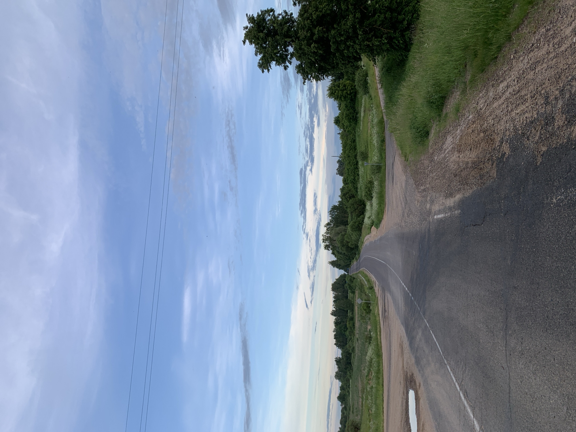 This image depicts a rural road intersection under a partially cloudy, light blue sky in Відзаўскі сельскі Савет, Belarus. The perspective is from a low angle on the main asphalt road, looking forward as it curves gently to the right and ascends slightly in the distance.
The sky occupies approximately the upper third of the frame, displaying various shades of light blue with scattered white and light grey stratocumulus clouds. Three dark, horizontal power lines traverse the sky in the upper-middle section. Small, dark specks, likely birds, are visible in the sky.
In the foreground, the asphalt main road exhibits signs of wear, including cracks and patches, with a continuous white line marking its center. A small, irregular puddle of water is present on the left edge of the road in the immediate foreground. To the right, a lighter-colored dirt and gravel road branches off, showing tire tracks. Another dirt area is visible on the left side of the main road.
The landscape surrounding the roads is characterized by lush green vegetation. Abundant grass, appearing as a mixture of cultivated and wild growth, lines the roadsides and fills extensive fields beyond. White wildflowers are intermingled with the grass in several areas. A dense collection of deciduous trees and shrubs forms a green band along the right side of the main road as it recedes, with a prominent, dark green tree standing on the far right. Further trees and vegetation form the horizon line across the mid-ground. A small, circular white road sign, indistinct in detail, is visible on the right side of the main road in the mid-distance. No human figures, animals, or vehicles are present in the scene.