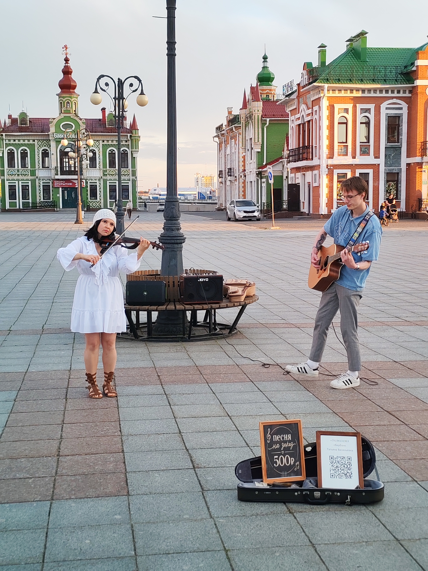 This media file captures a street performance in Yoshkar-Ola, Russia. Two musicians are actively playing on a paved square in front of colorful, ornately decorated buildings. On the left, a woman in a white dress and a white headscarf plays the violin, her head tilted as she concentrates on her music. She wears brown gladiator-style sandals. To her right, a young man in a denim shirt and gray pants plays an acoustic guitar. He wears glasses and white sneakers.
Between them, a wooden bench holds two amplifiers, one labeled "JOYO". In the foreground, an open violin case displays two framed signs. One sign, a chalkboard, has musical notes and text that reads "песня на заказ 500P" (song by order 500 rubles). The second sign provides a phone number "+79194131963" with "сбербанк" (Sberbank) and a name "Татьяна Васильевна" (Tatiana Vasilievna), along with a QR code.
The setting appears to be a public square, possibly a main street, given the style of the buildings and the presence of vehicles in the background. The lighting suggests it's either early morning or late afternoon, with the golden hue of the sun casting long shadows. The sky is a soft, hazy blue, indicating fair weather. The overall mood is one of artistic engagement and a pleasant urban atmosphere. The architecture of the buildings, with their vibrant colors and intricate detailing, is a prominent feature of the scene, hinting at a location with distinctive cultural or historical significance.
