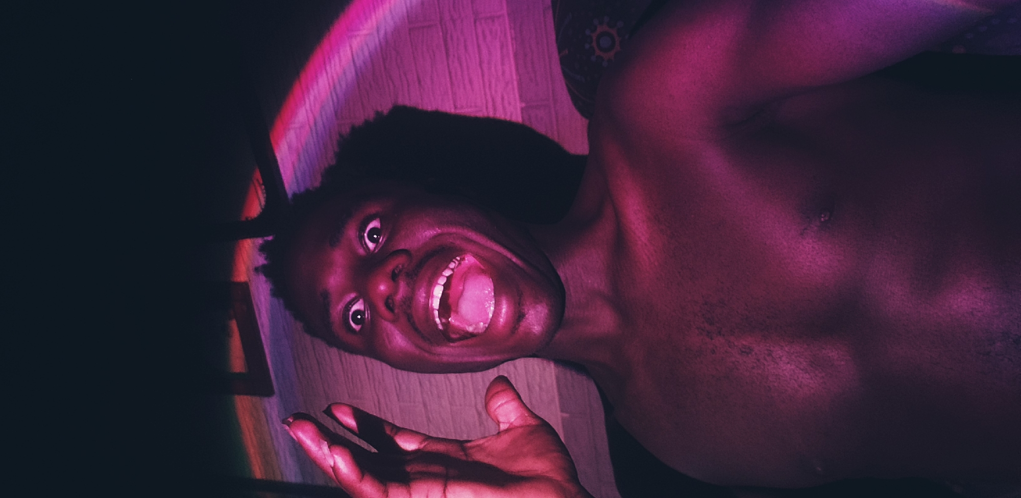 A young Black male, shirtless, is captured in a close-up, low-angle shot. He has dark skin and appears to be looking directly at the camera with wide eyes and an open mouth, as if shouting or exclaiming. His left hand is raised and open, close to his face. The lighting in the scene is dramatic, with vibrant pink and purple hues casting shadows and highlighting his features and torso. A rainbow-like light effect arcs across the upper right portion of the image. The background appears to be a brick wall, with some patterned fabric visible on the right. The image's overall tone is energetic and possibly playful due to the subject's expression and the colorful lighting. The presence of a brick wall suggests an indoor or semi-enclosed space.