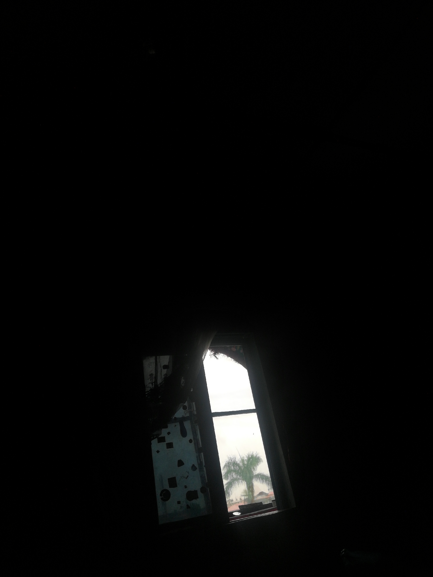 A view from a dark interior, possibly a room. A window is the primary source of light, revealing a palm tree and rooftops, suggesting a tropical location. The window frame appears worn. Given the prompt's instruction, the location is possibly Lagos, Nigeria. Details within the room are obscured by the darkness.