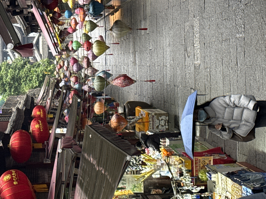 The image depicts an uphill, stone-paved street within a market area in Pengshui Miao and Tujia Autonomous County, China. Numerous lanterns are strung overhead; these include spherical red lanterns with yellow tassels displaying Chinese characters and a canopy of multicolored, teardrop-shaped fabric lanterns with red tassels. Traditional Chinese architectural elements, including dark-tiled roofs and wooden structures, line both sides of the street, with dark awnings extending over various shop fronts.
In the mid-foreground, a female subject is walking uphill, holding a light blue umbrella. She wears a light-colored puffer jacket, dark trousers, dark gloves, and a light blue surgical mask. A dark shoulder bag is carried on her right side. To her left, at a market stall, another individual in a dark puffer jacket is visible, appearing to tend to goods that include fresh fruit and items on sticks. Further up the street, additional indistinct figures are present amidst various stalls displaying merchandise and signage with Chinese characters, one prominent sign reading "甘蔗汁" (sugarcane juice). The street surface is composed of grey, rectangular stone blocks. Green foliage is visible in the upper background, indicating a natural element beyond the immediate structures.