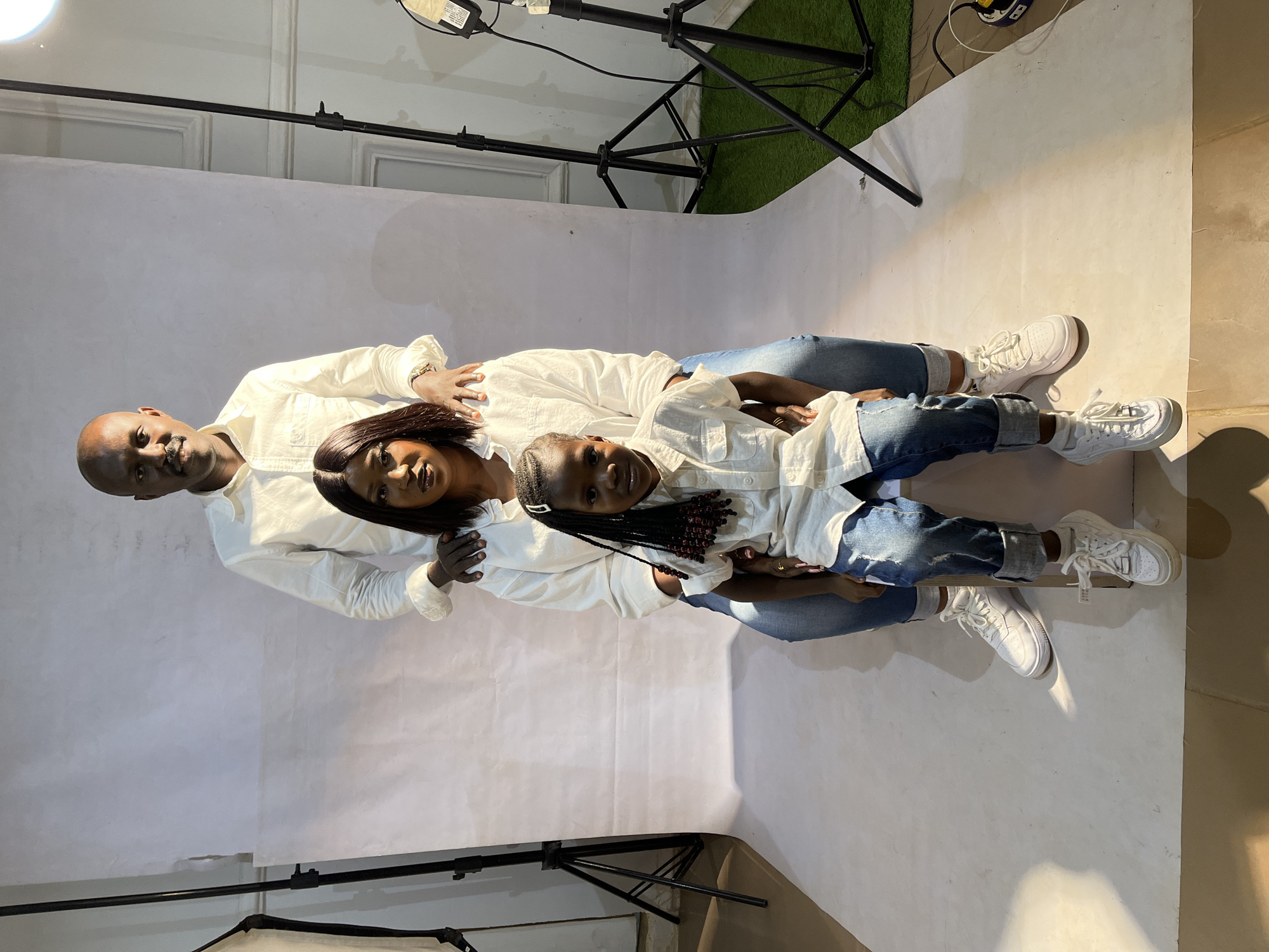 A photograph shows a family of three posing in a studio setting. The father, mother, and daughter are lying on a white backdrop, all facing the camera. The father is at the top of the frame, with his left hand resting on the mother's shoulder. The mother is in the middle, and the daughter is positioned in front of her. All three are wearing white shirts and blue jeans. They all have white sneakers on.
A lighting setup is visible in the background, including a stand with a light and a power strip. A patch of artificial green grass can also be seen in the upper right corner. The photograph appears to have been taken at a studio in Barakin Akawo, Nigeria.