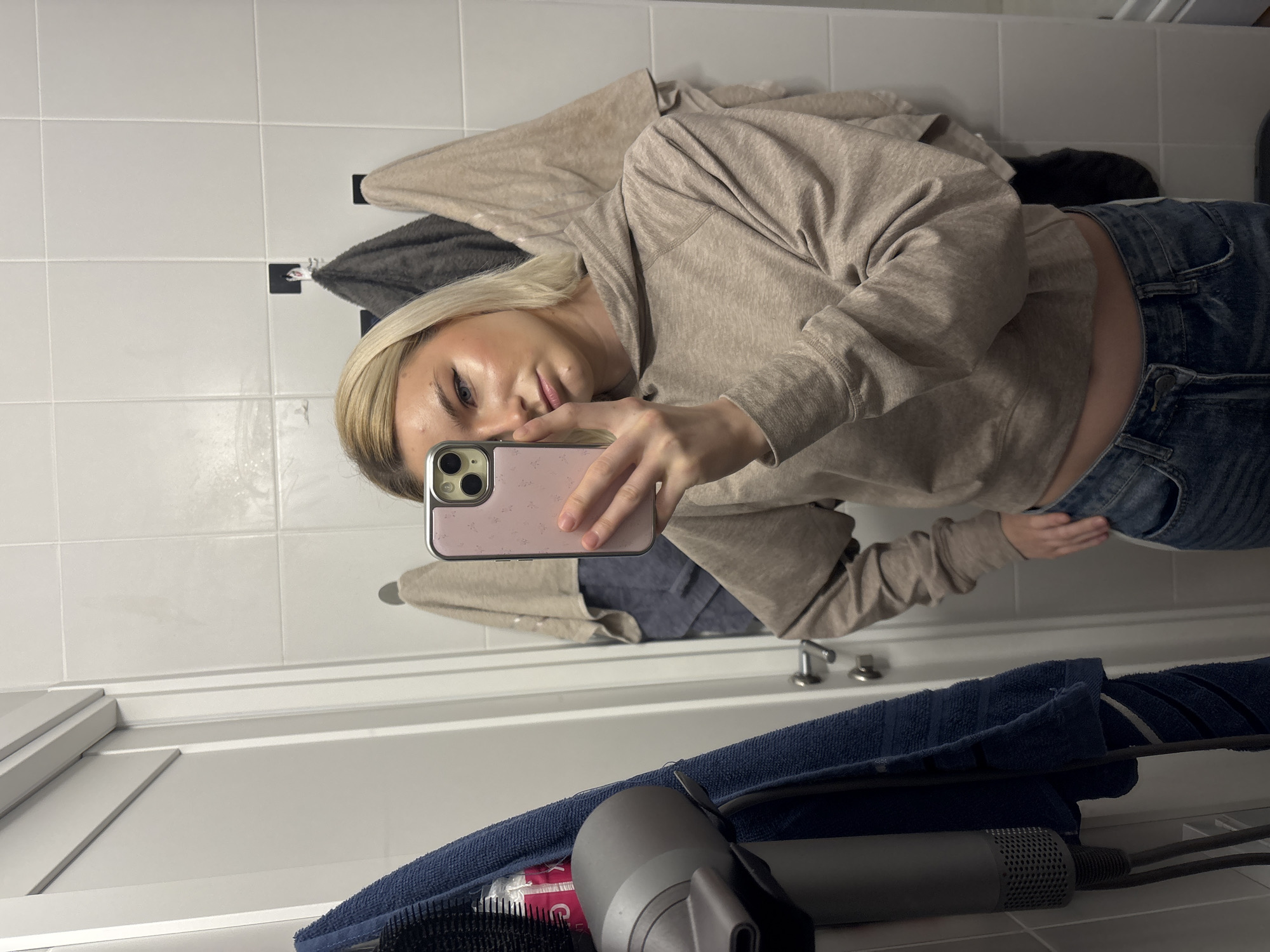 A mirror selfie, showing a woman with blond hair wearing a beige hoodie and blue jeans. She is holding a phone with a pink case. Behind her are white tiled walls and some towels hanging on hooks. A blue towel and a hairdryer are visible in the foreground. The location is a bathroom. No information about a city or Russia is available in the picture.