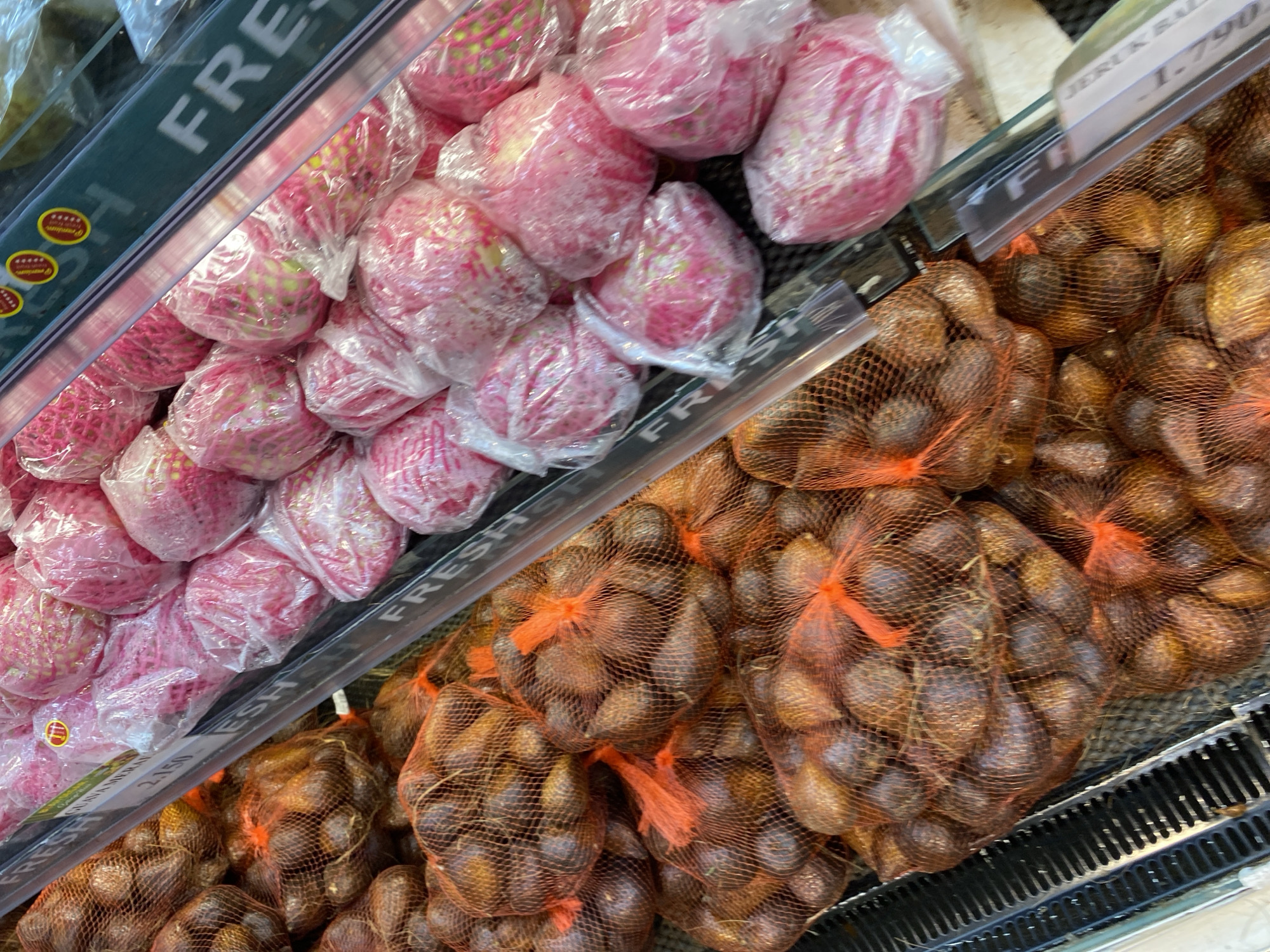 The image shows a close-up view of fruit displayed in a refrigerated section of a supermarket. On the left, there are several bags filled with brown, oval-shaped fruits, likely salak, each tied with orange netting. These bags are stacked on a black grated shelf. To the right of the salak, there are rows of pink fruits, possibly guavas or apples, each individually wrapped in clear plastic, some with pink netting. A green price tag with white text is visible, with the word "FRESH" partially showing, and below it, "GUAVA MERAH" and "2.150". Another price tag with "JERUK BALI" is visible on the right. The overall environment is that of a brightly lit retail space. The specific location within the city of Banda Aceh, Indonesia, cannot be determined from the image alone.