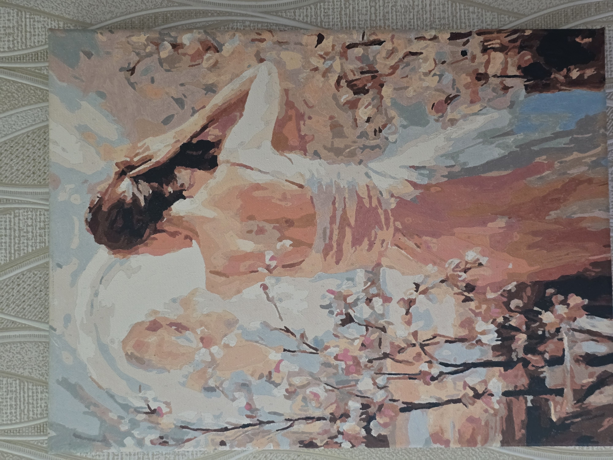 The image shows a painting of a person lying down, possibly on a bed or in a field of flowers, with a white cloth draped over them. The painting uses soft, pastel colors, with shades of pink, peach, and light blue dominating the scene. The person's hair is dark, and their face is partially obscured. There are also elements that suggest a natural setting, such as branches with small pink and white flowers, resembling cherry blossoms. The overall impression is one of tranquility and gentle beauty. The painting is rendered in a style that appears to be paint-by-numbers, with distinct sections of color that suggest the artist followed a pre-drawn outline. The painting is set against a textured background, possibly a wallpaper or fabric. There is no visible text in the image.