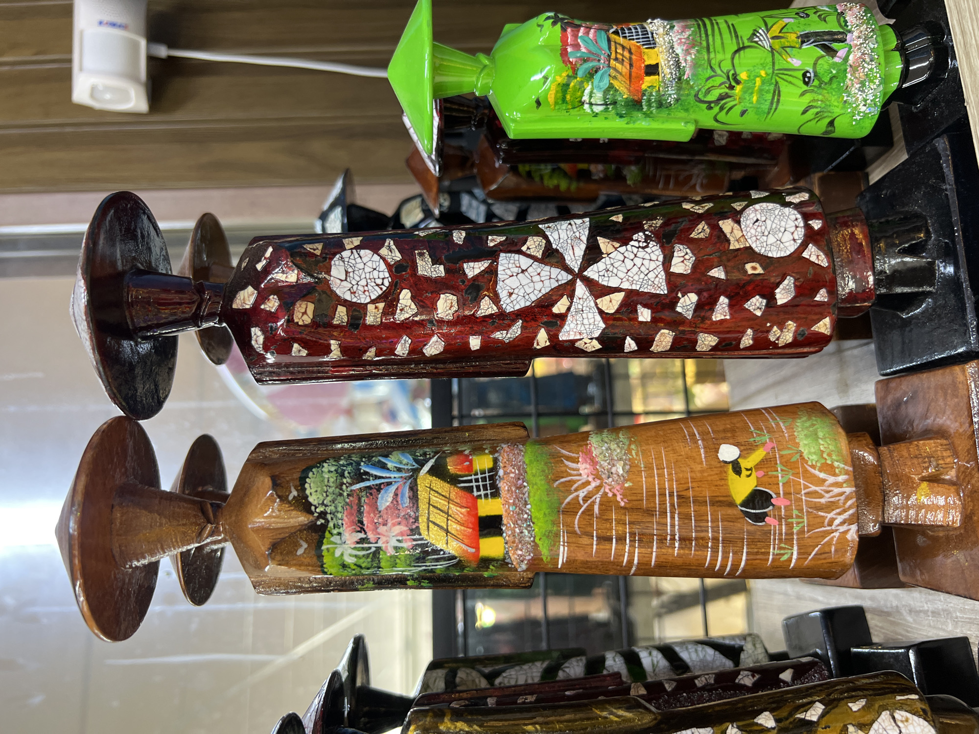 The image shows a display of wooden souvenir figures, likely in a shop in Nha Trang, Vietnam. The figures are tall and slender, with conical hats. They are decorated with painted scenes depicting rural Vietnamese life, including houses, rice paddies, and people working. The figure on the left is made of natural wood and features a colorful depiction of a house with a thatched roof, palm trees, and a person tending to crops. The figure in the center is dark red and decorated with a mosaic-like pattern of white and earth-toned fragments. The figure on the right is bright green and also painted with a rural scene, including a house, vegetation, and a person wearing a conical hat. Several other figures are visible in the background, some partially obscured. The figures are standing on black bases, and the display appears to be on a shelf or counter. The background shows shelves with other items and a glimpse of what might be a window or glass partition. A white electronic device with a cable is visible on the right side of the image, mounted on a wooden surface.