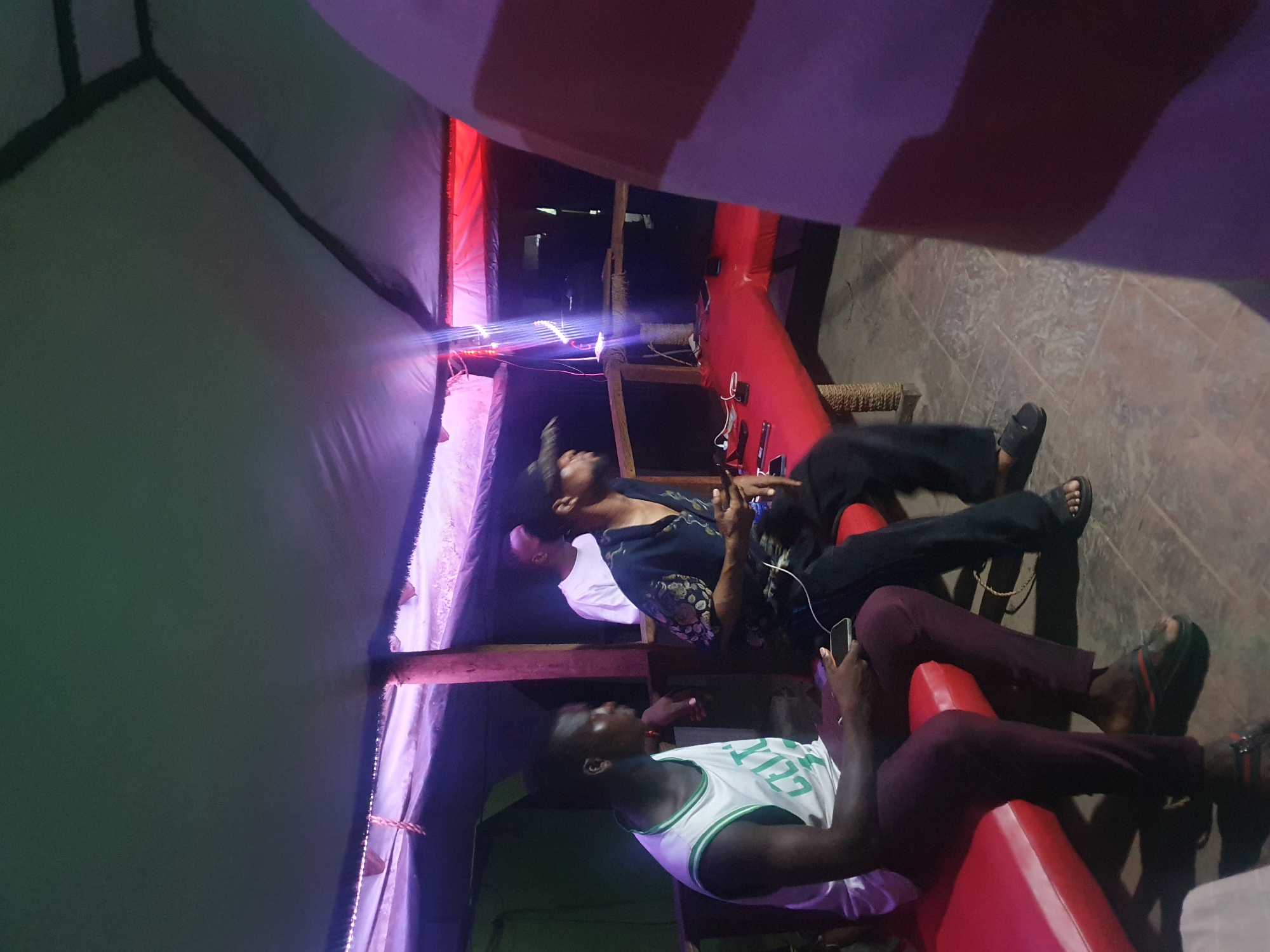 Two individuals are seated indoors, possibly in a restaurant or informal gathering place within Lokoja, Nigeria. A man wears a white Boston Celtics tank top and maroon-colored pants. A second individual sits above and behind, wearing a patterned short-sleeved shirt and dark pants. Both are using smartphones. A red and white striped fabric drapes from the upper part of the image. A light fixture shines above. A green and grey fabric structure is visible on one side. Mobile phones are charging on a red structure. The floor is tiled.