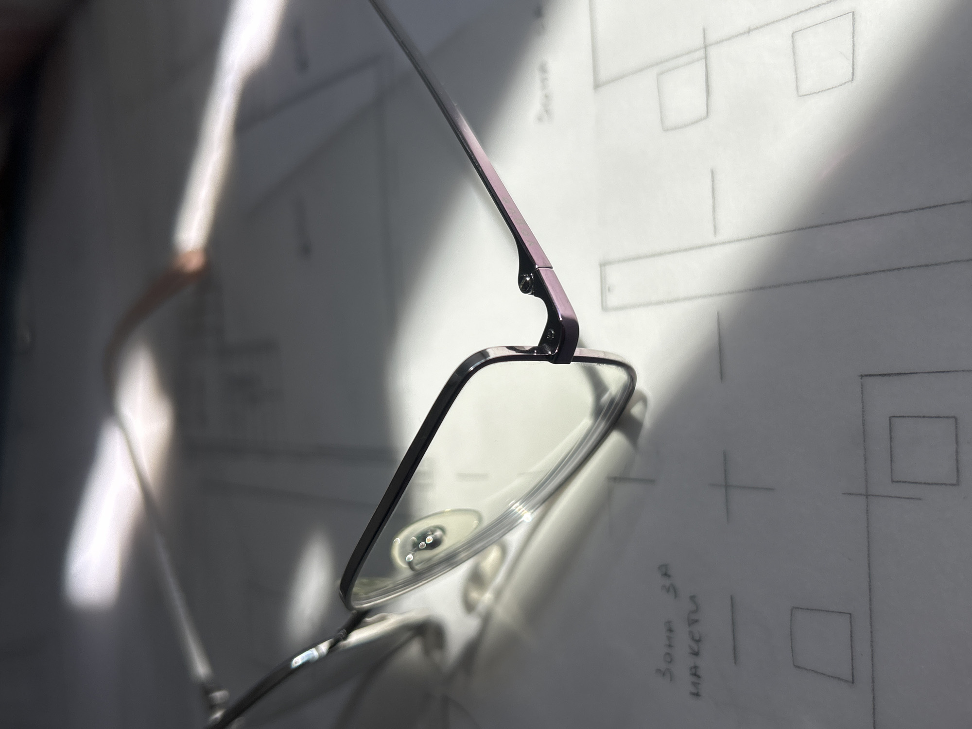 The image shows a pair of eyeglasses resting on a white piece of paper with technical drawings or diagrams. The frames of the glasses are dark colored with a contrasting pinkish hue on the temples. The paper has various geometric shapes drawn on it, including squares and lines, with some text in what appears to be Bulgarian. The lighting is uneven, creating strong shadows and highlights across the surface, possibly from a nearby window. There is not enough information to determine the location in Plovdiv, Bulgaria.