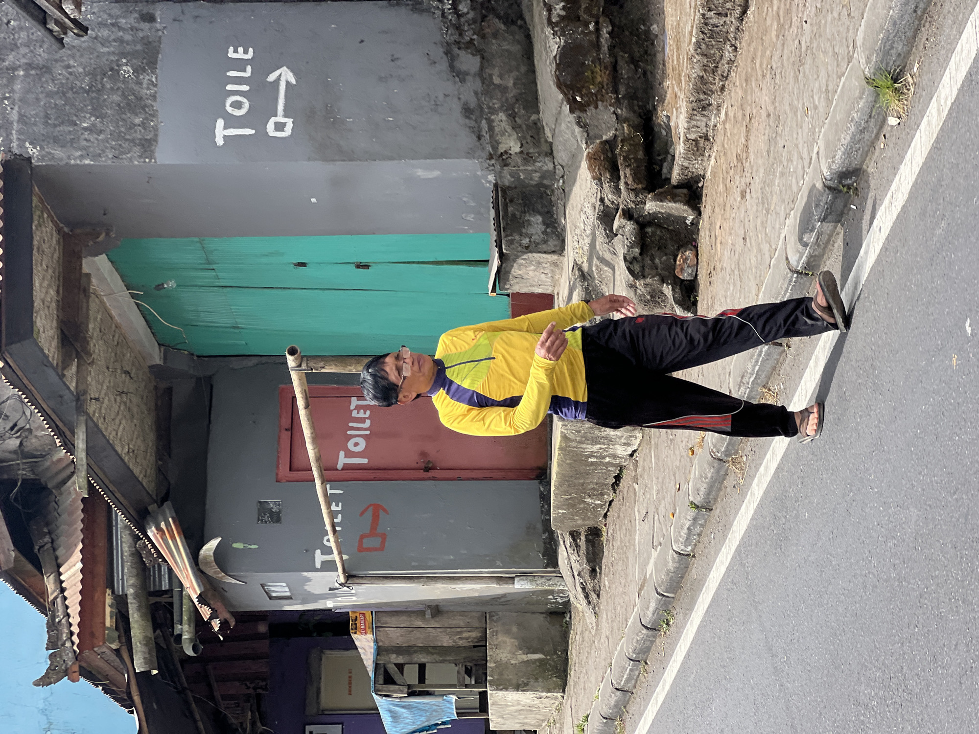 A person walks on a sidewalk next to a street in Kintamani, Indonesia. The person is wearing a yellow and purple long-sleeved shirt and black pants with red stripes. They are also wearing sandals. The street is paved and has a white line painted on the right side. The sidewalk is made of concrete and has grass growing in some cracks. Behind the sidewalk are several buildings with weathered facades. One building has a green door and another has a red door with "TOILET" painted on it in white. There are also signs with arrows pointing to the right, indicating the direction of the toilets. The sky is visible at the top of the image and is blue.