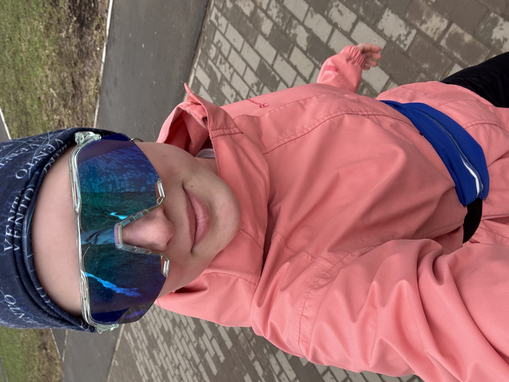 A person wearing a pink jacket, a blue headband with text, and reflective blue sunglasses is posing outdoors. The person is also wearing a blue fanny pack. The background appears to be a paved area with brick-like patterns. To the left, there's a patch of grass. The photo was likely taken in Chaikovsky, Russia.