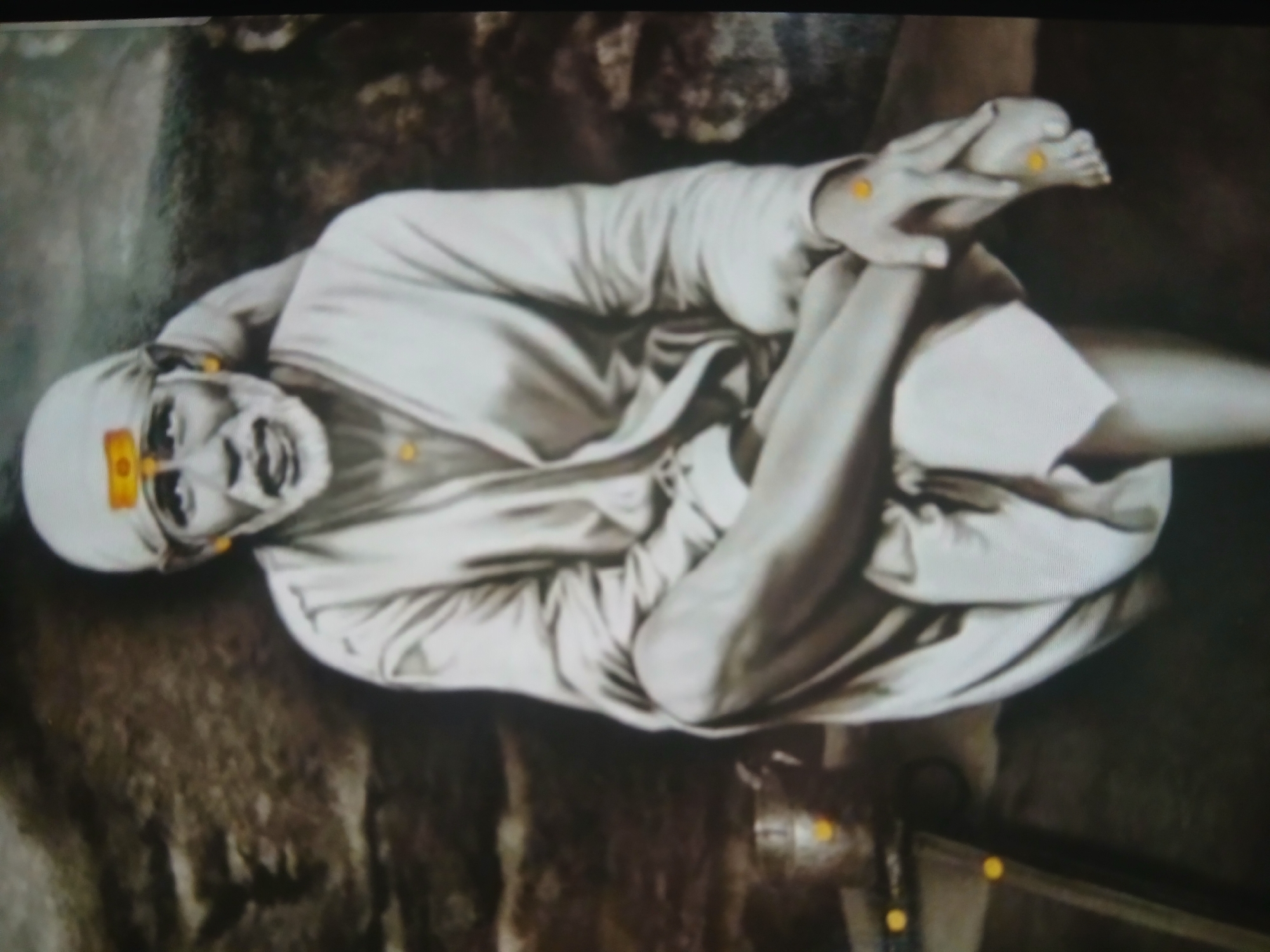 A color image shows a portrait of Sai Baba sitting cross-legged. He wears a white robe and a head covering. He has a tilak on his forehead. His right hand holds his left foot. He is surrounded by a dark, blurry background. I am unable to provide the location as Bengaluru because there is no indication of this within the image.