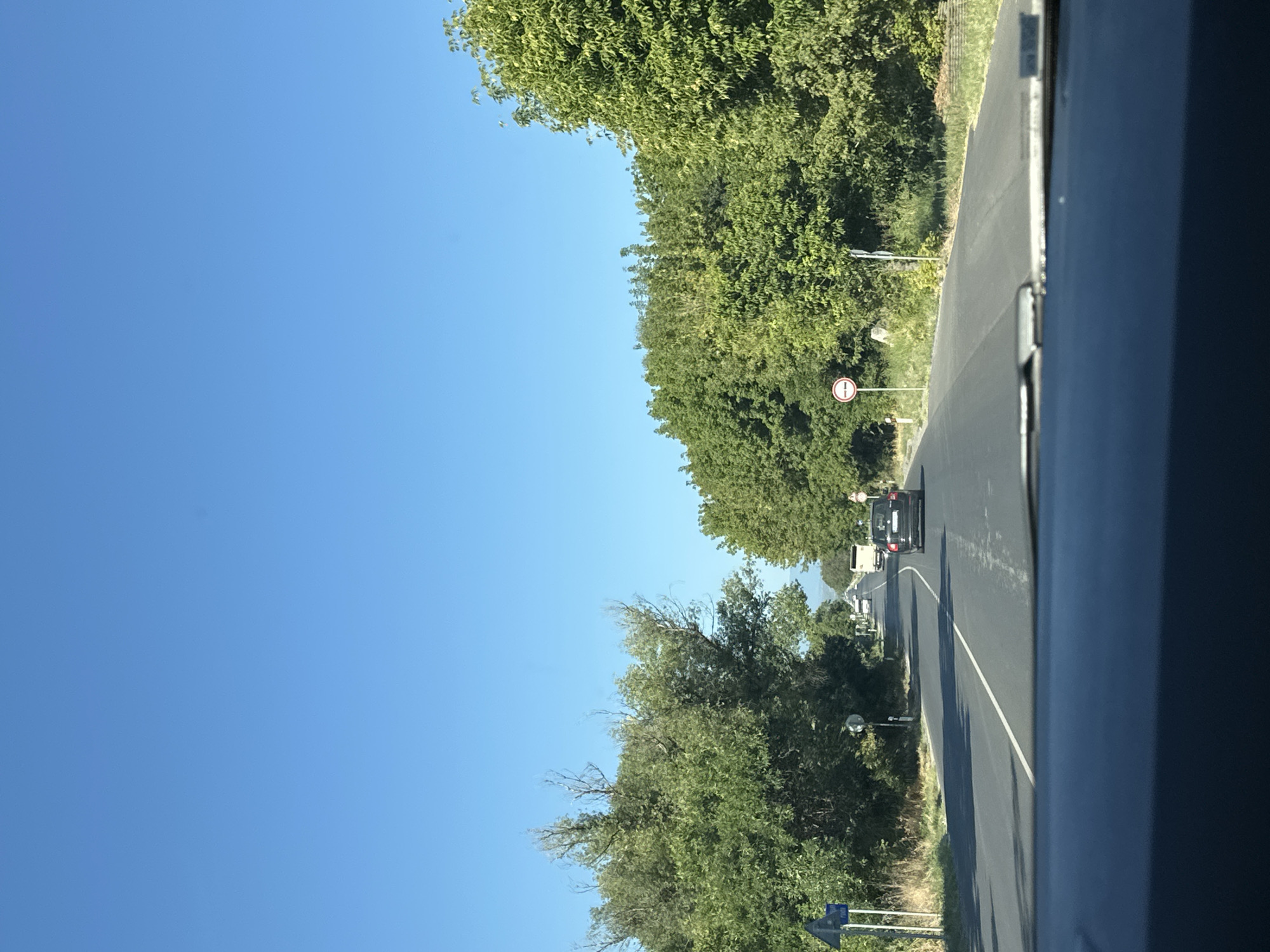 The image depicts a view from a vehicle traveling on a two-lane asphalt road lined with dense green trees on both sides. The sky is clear and blue. Several vehicles are visible on the road ahead, including a white bus and a dark-colored car. There are also road signs on the side of the road. The location is specified as the city of Kadievo, Bulgaria. The overall scene suggests a sunny day on a rural or suburban road.