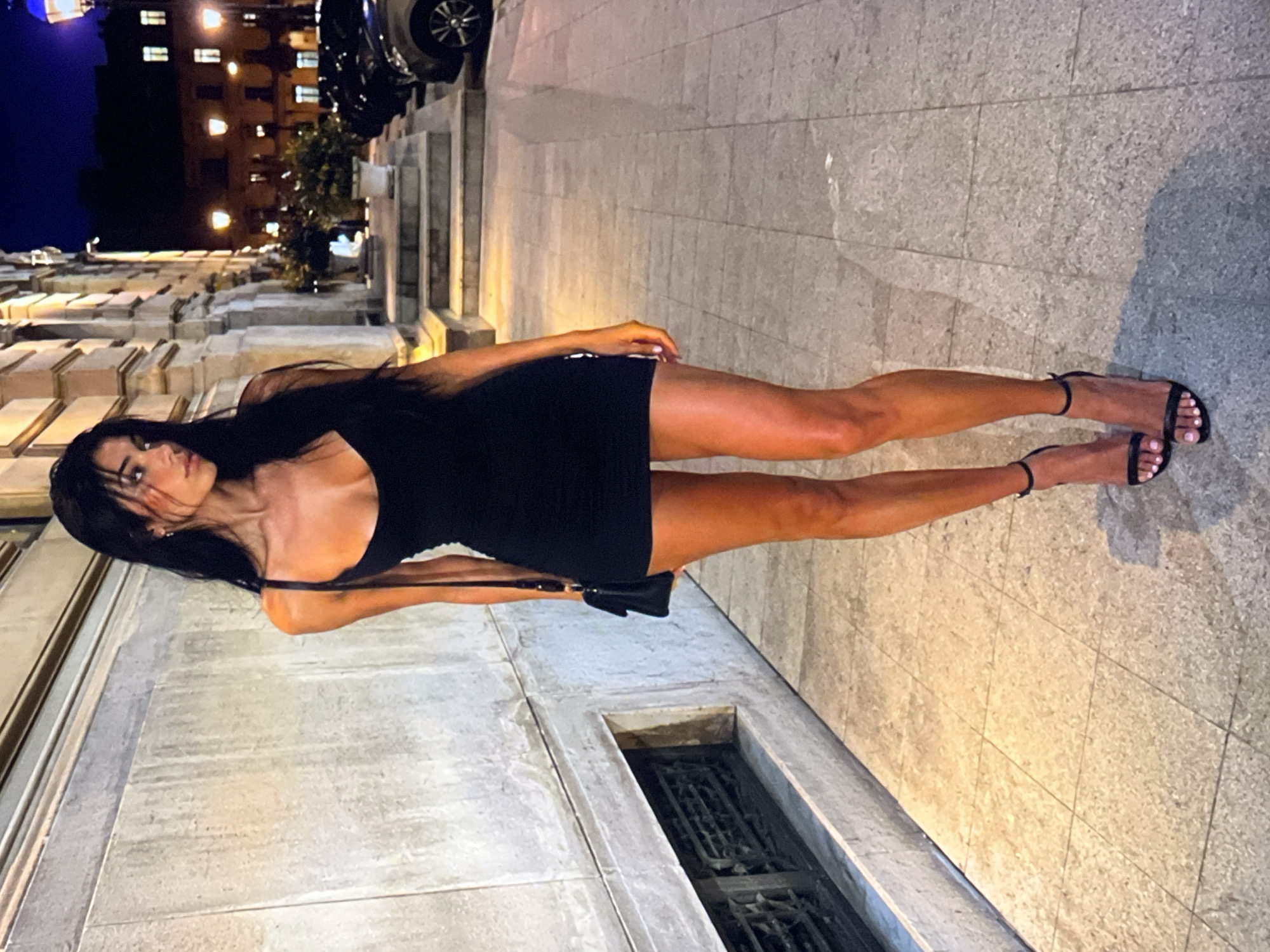 A full-body, outdoor, night-time medium shot captures a young, slender woman with long, dark hair standing on a tiled sidewalk in front of a building with stone architectural details. She is wearing a form-fitting black mini dress with thin straps and black stiletto sandals. A small black purse hangs from her right shoulder. The background shows a street with parked cars, illuminated windows, and streetlights. The lighting is dim, with artificial lights casting a warm glow on the scene.
