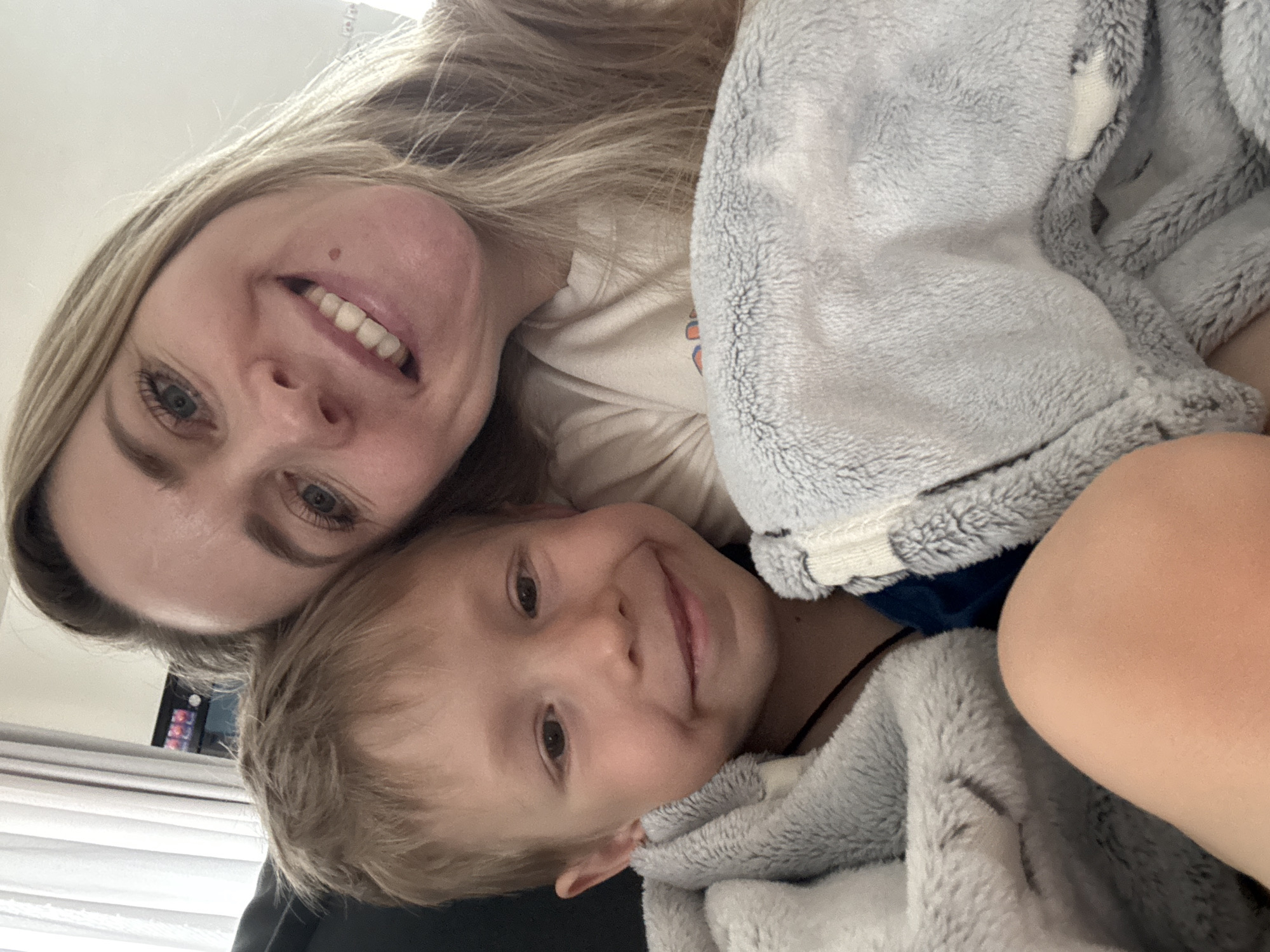A selfie image shows a woman and a young boy huddled together closely. The woman, positioned at the top of the frame, has fair skin, blonde hair, and light-colored eyes. She is smiling broadly, revealing her teeth, and has a mole near her mouth. The boy, located below her, also has fair skin and blonde hair. He is smiling gently. Both are partially covered in a light gray, fuzzy hooded blanket. The image appears to be taken indoors with some blurred background elements visible. The location cannot be determined as Chaikovsky, Russia, based on the available image content.