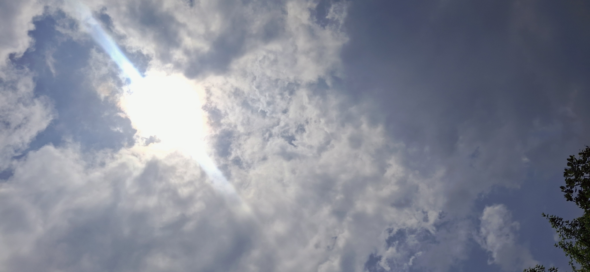 A view of the sky over Vallecito, Venezuela, shows a bright sun partially obscured by clouds. A sunbeam emanates from the sun, casting a diagonal ray of light across the frame. The clouds are a mix of white and gray, with some appearing wispy and others more dense. In the bottom left corner of the image, the tops of green trees are visible against the sky. The overall impression is of a slightly overcast but bright day.
