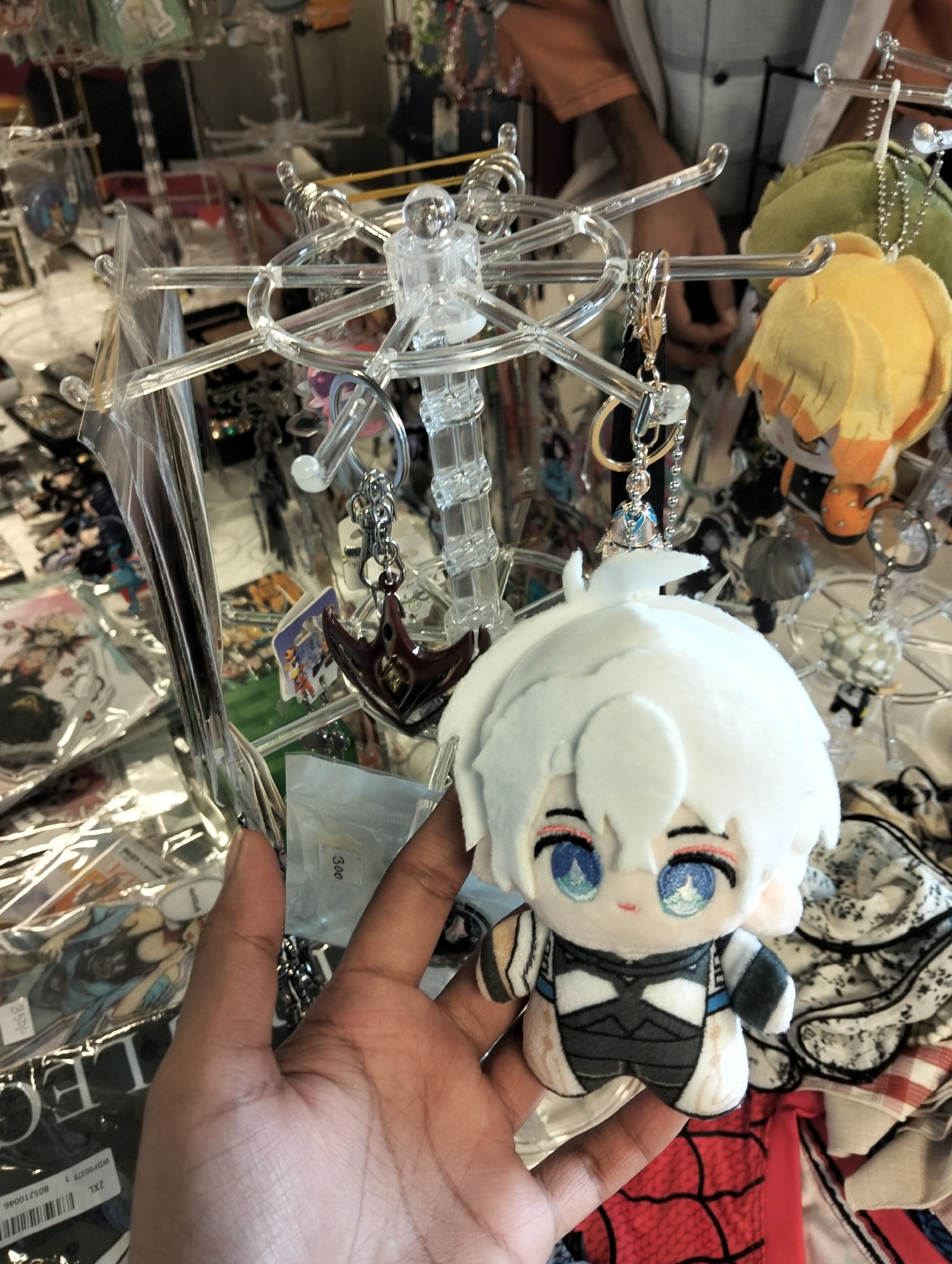 A hand holds a plush toy with white hair and blue eyes in front of a display of various merchandise. The background appears to be a stall or shop with many items hanging on clear plastic racks, including keychains and other small accessories. The lighting is bright, suggesting an indoor setting, possibly a market or store. The overall scene captures a close-up view of the plush toy and the hand holding it, with a blurred background of merchandise. There is no visible indication of the specific location within Dhaka, Bangladesh.
