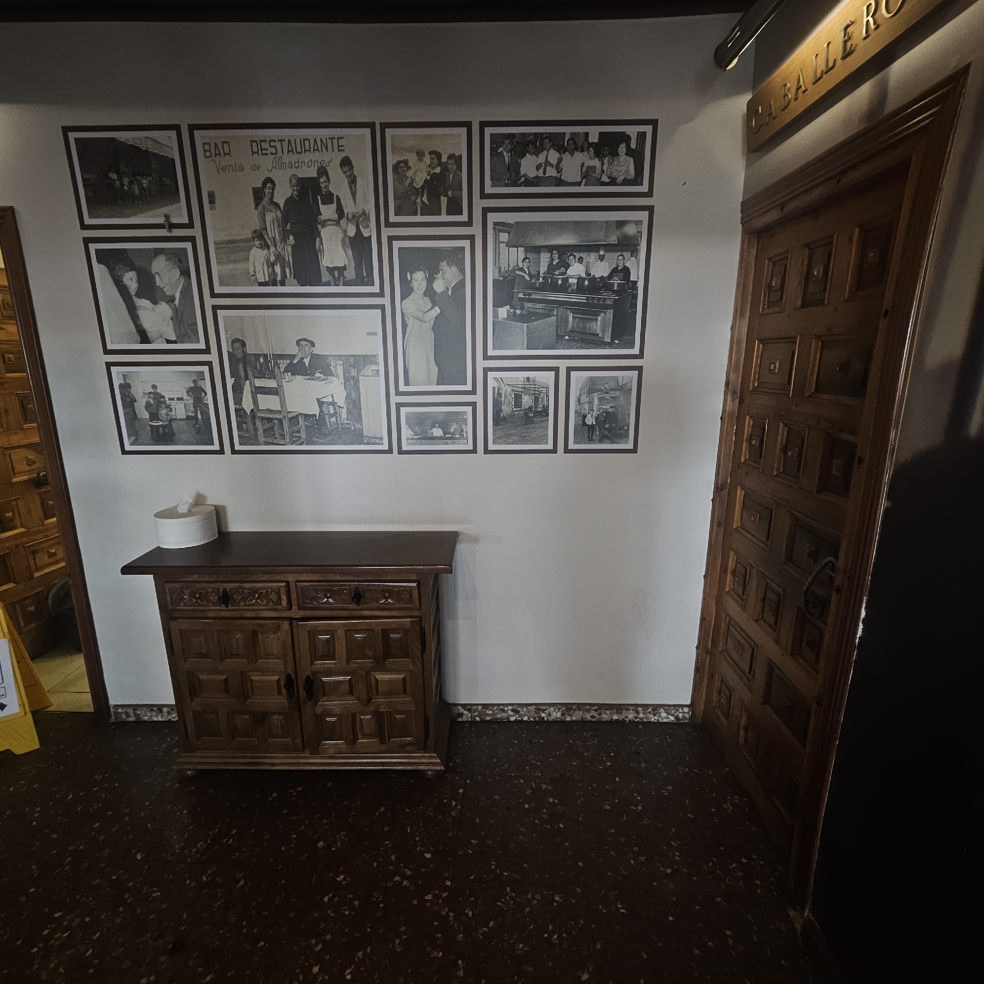 The image shows an interior scene with a wall adorned with framed black and white photographs. The photographs display various scenes, including people, groups, and what appear to be historical moments, with one picture featuring text that reads "BAR RESTAURANTE VENTA de Almadrones". A wooden cabinet with carved details is positioned below the photographs. To the right, a large wooden door with intricate paneling and a rope handle is visible, along with a sign above it that reads "CABALLERO" in gold lettering. The floor is dark and speckled.