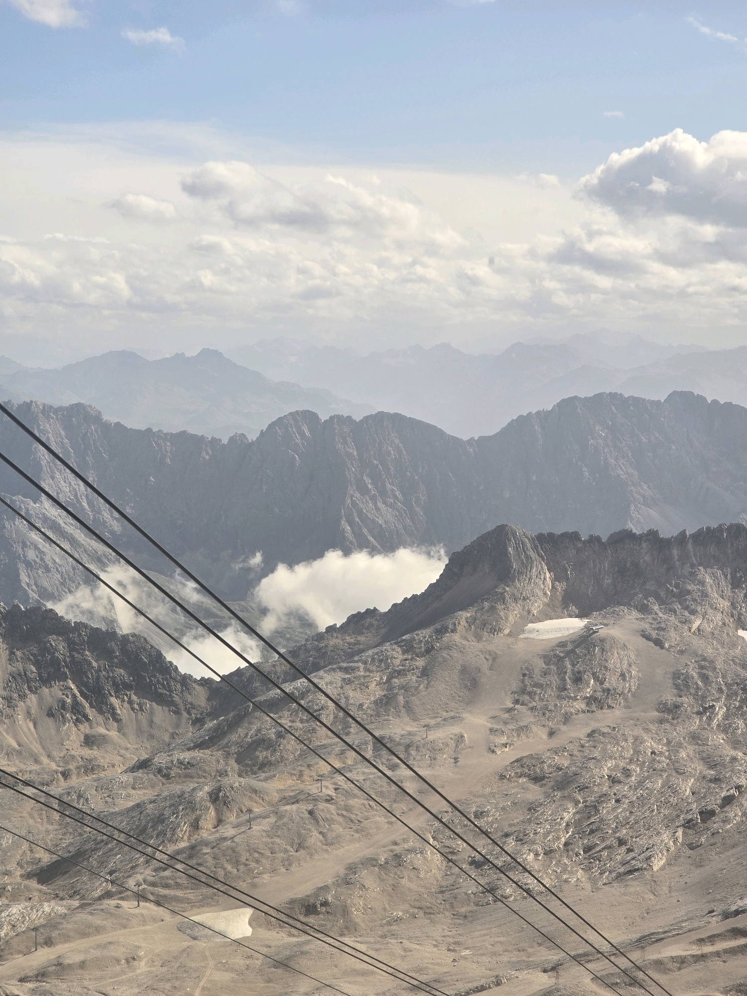 This image captures a mountainous landscape with layers of peaks receding into a hazy sky. In the foreground, several dark cable car wires diagonally traverse the scene, suggesting a transportation system. The terrain is rugged and rocky, with patches of snow or ice visible on some slopes. In the middle ground, clouds are nestled within the valleys between mountains, adding a sense of depth. The overall impression is one of a high-altitude, remote natural environment. The location is identified as Grainau, Germany, likely in the Bavarian Alps, famous for its mountainous terrain and cable car access to peaks like the Zugspitze.